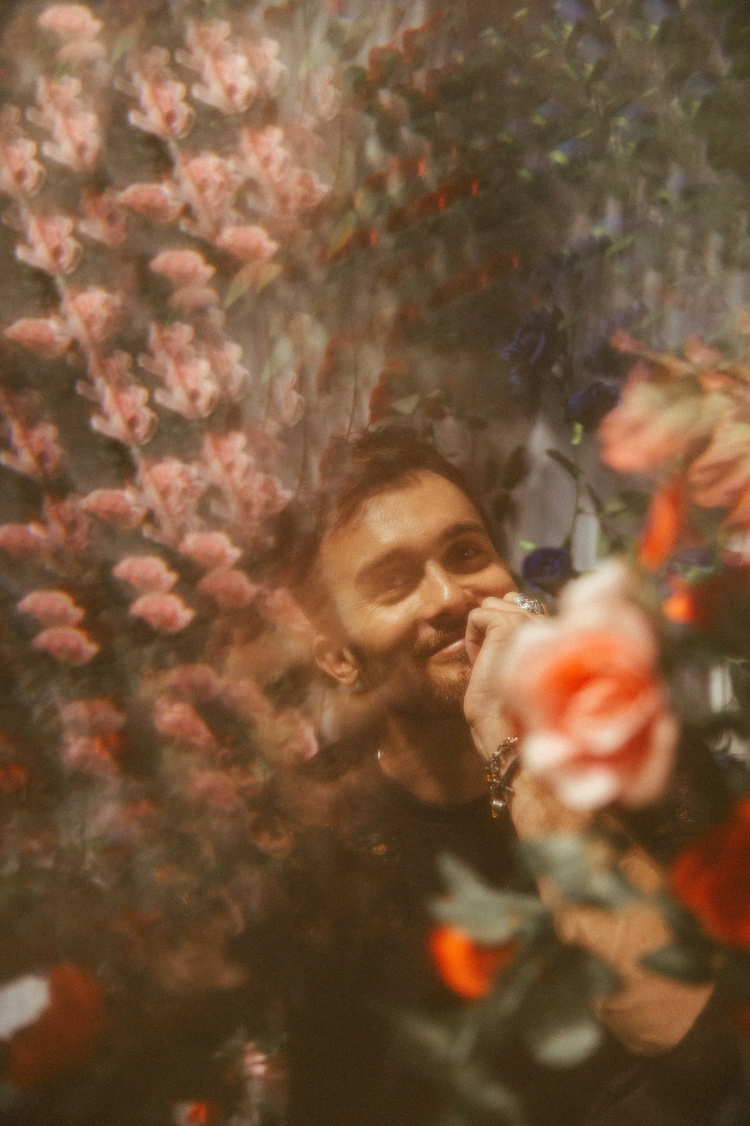 A man smiling and resting his chin on his hand surrounded by a dense display of pink and purple flowers.