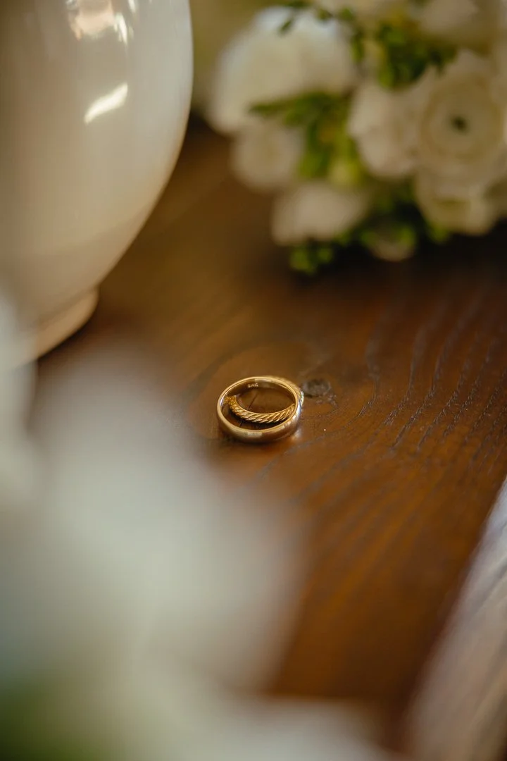 Close-up of a pair of gold wedding rings on a wooden surface, with blurred white flowers and a beige vase in the background.