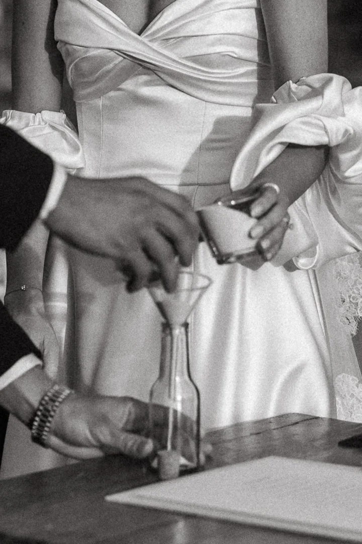 Black and white photo of a bride in a wedding dress holding a small mug, standing at a table with a bottle and a vase, with another person's hands pouring a drink into the mug.