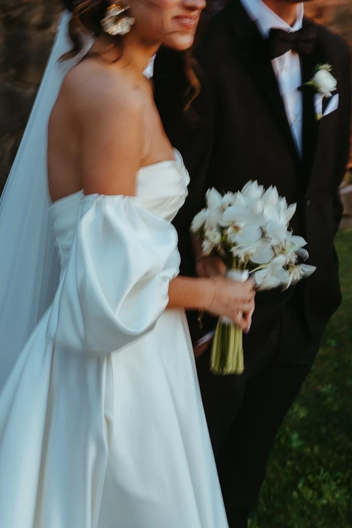 Bride in a white wedding dress holding a bouquet of white flowers walking beside a groom in a black tuxedo with a white boutonniere.