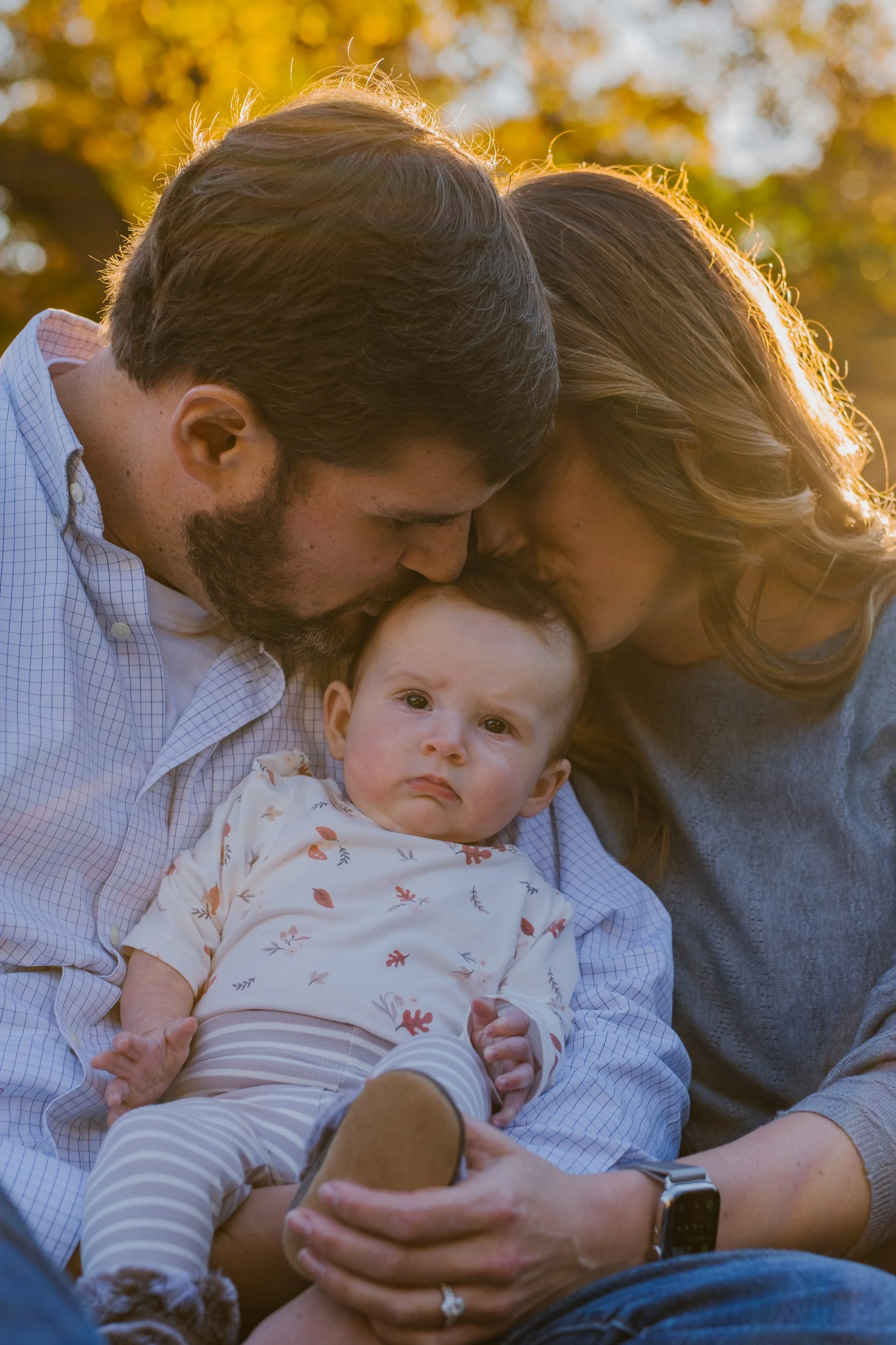 A couple kisses their baby on the forehead in an outdoor setting with golden trees in the background.