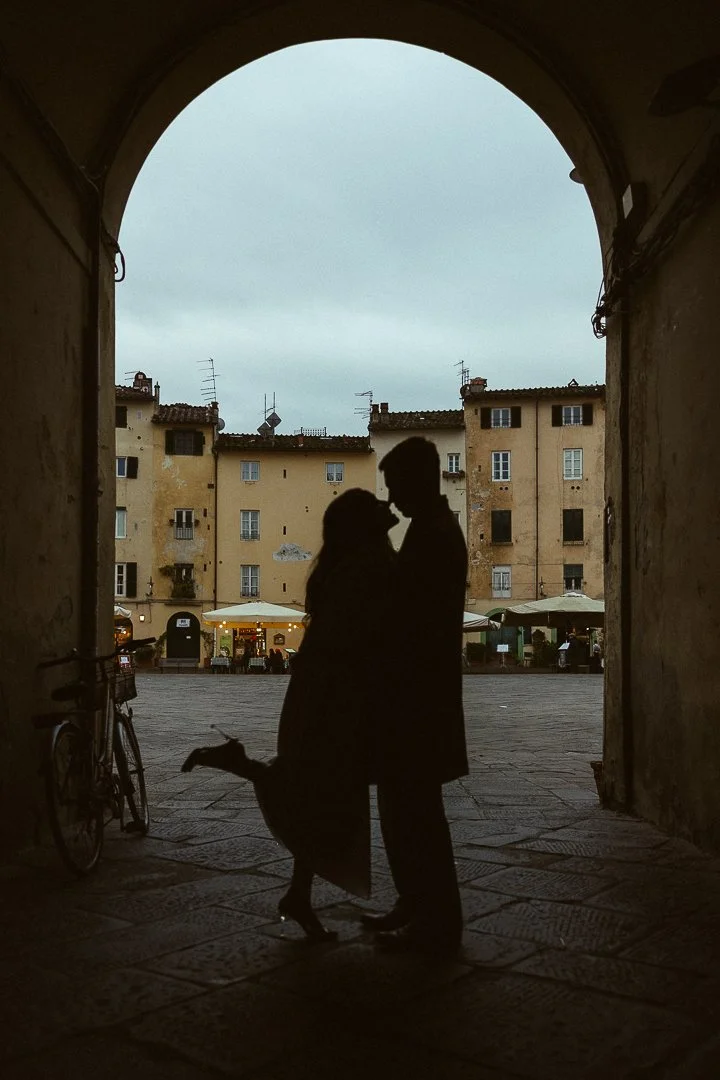 Silhouette of a couple kissing under an archway with historic buildings in the background during dusk.