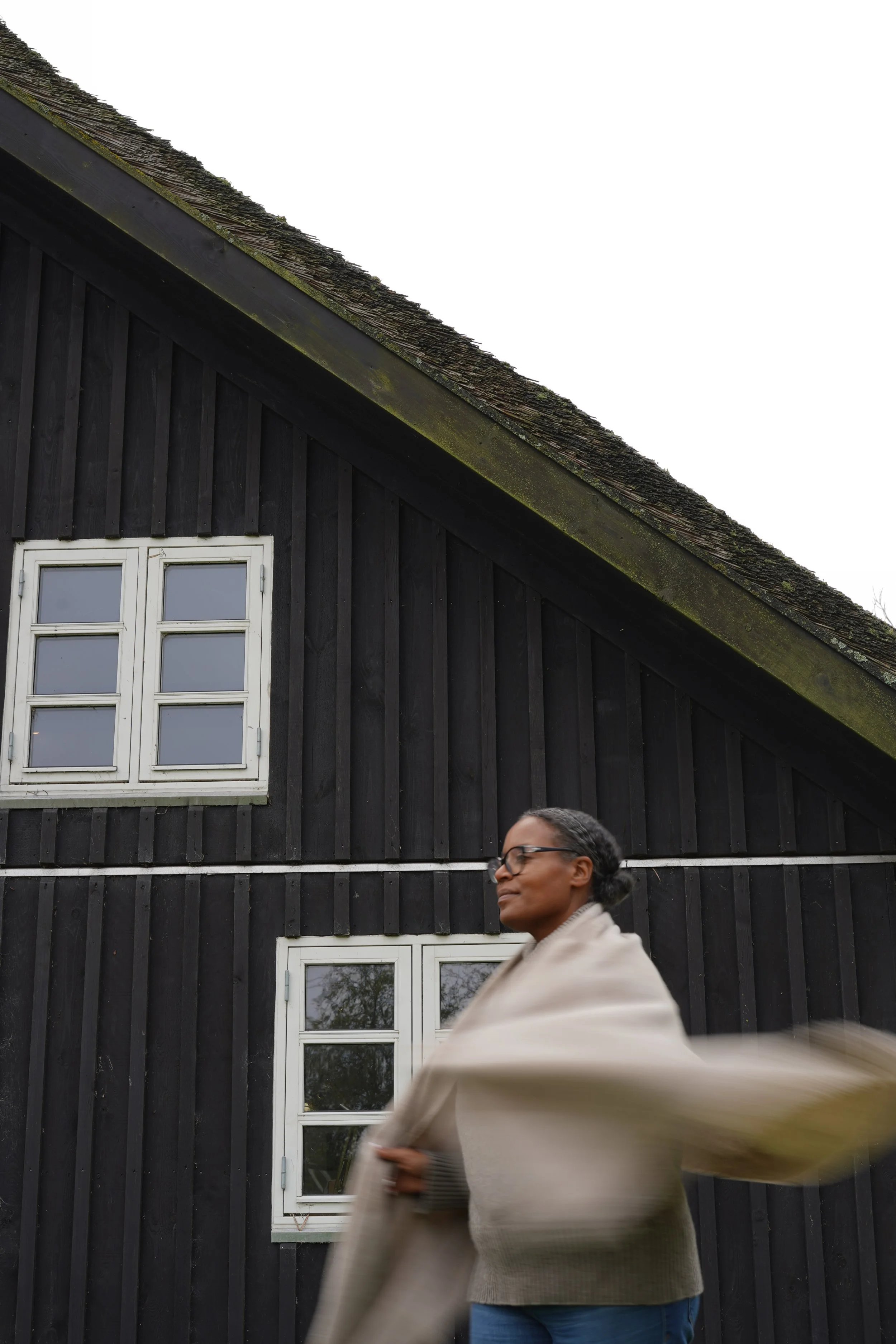 A woman with glasses wearing a light beige coat standing in front of a black wooden house with white window frames. She appears to be adjusting her coat or scarf, and she is slightly blurred due to motion.