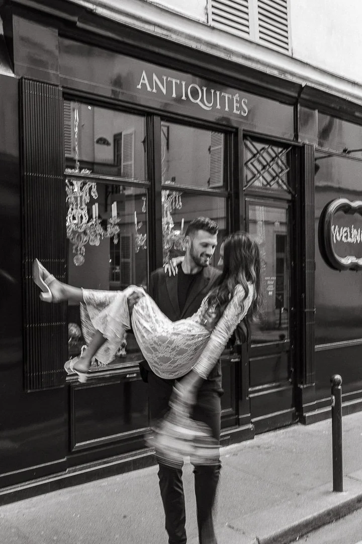 A man carrying a woman in his arms in front of an antique shop window, with chandeliers visible inside, in black and white.