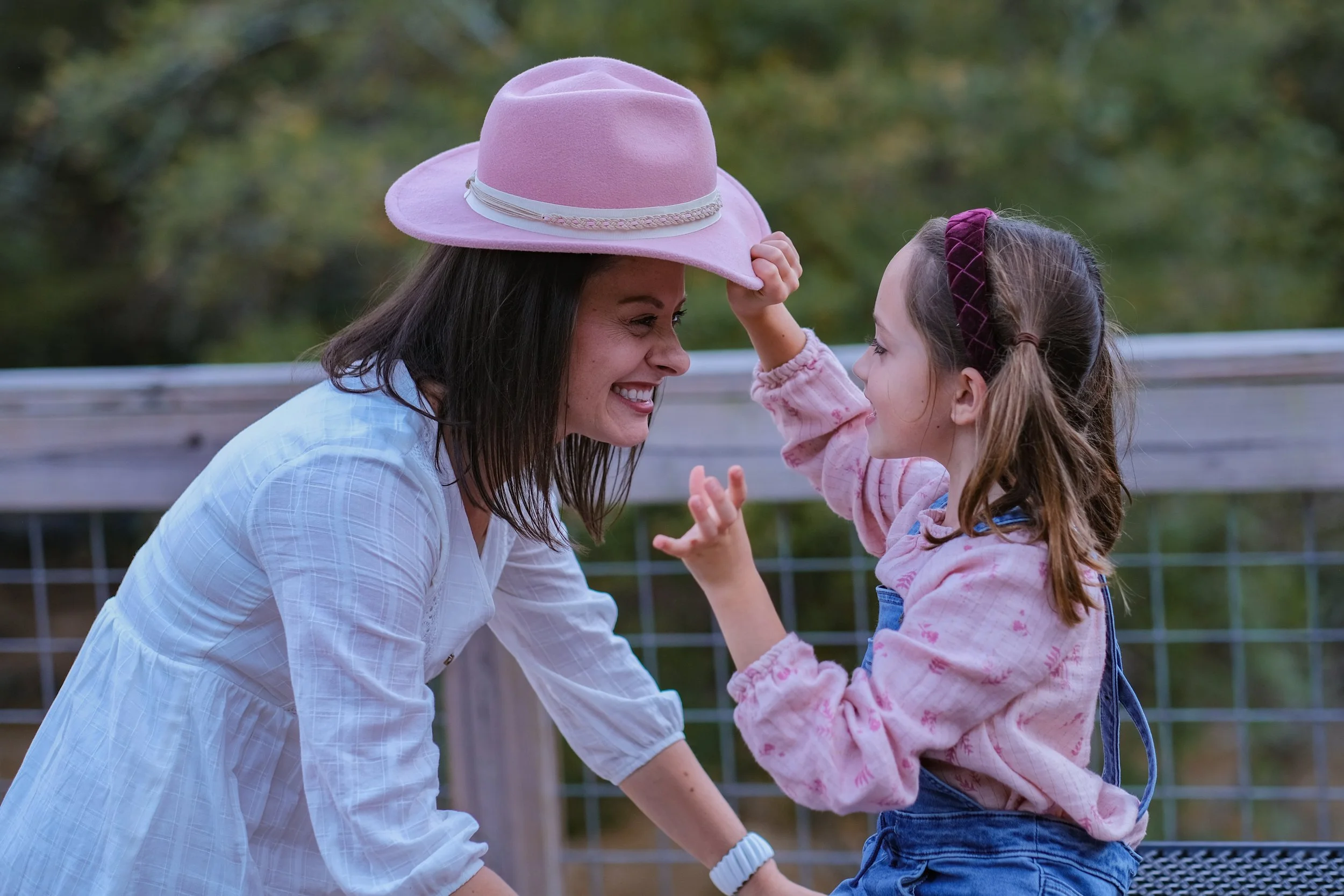 A woman and a young girl smiling and touching foreheads outside, with the woman wearing a pink hat and the girl wearing a headband and pink sweater.
