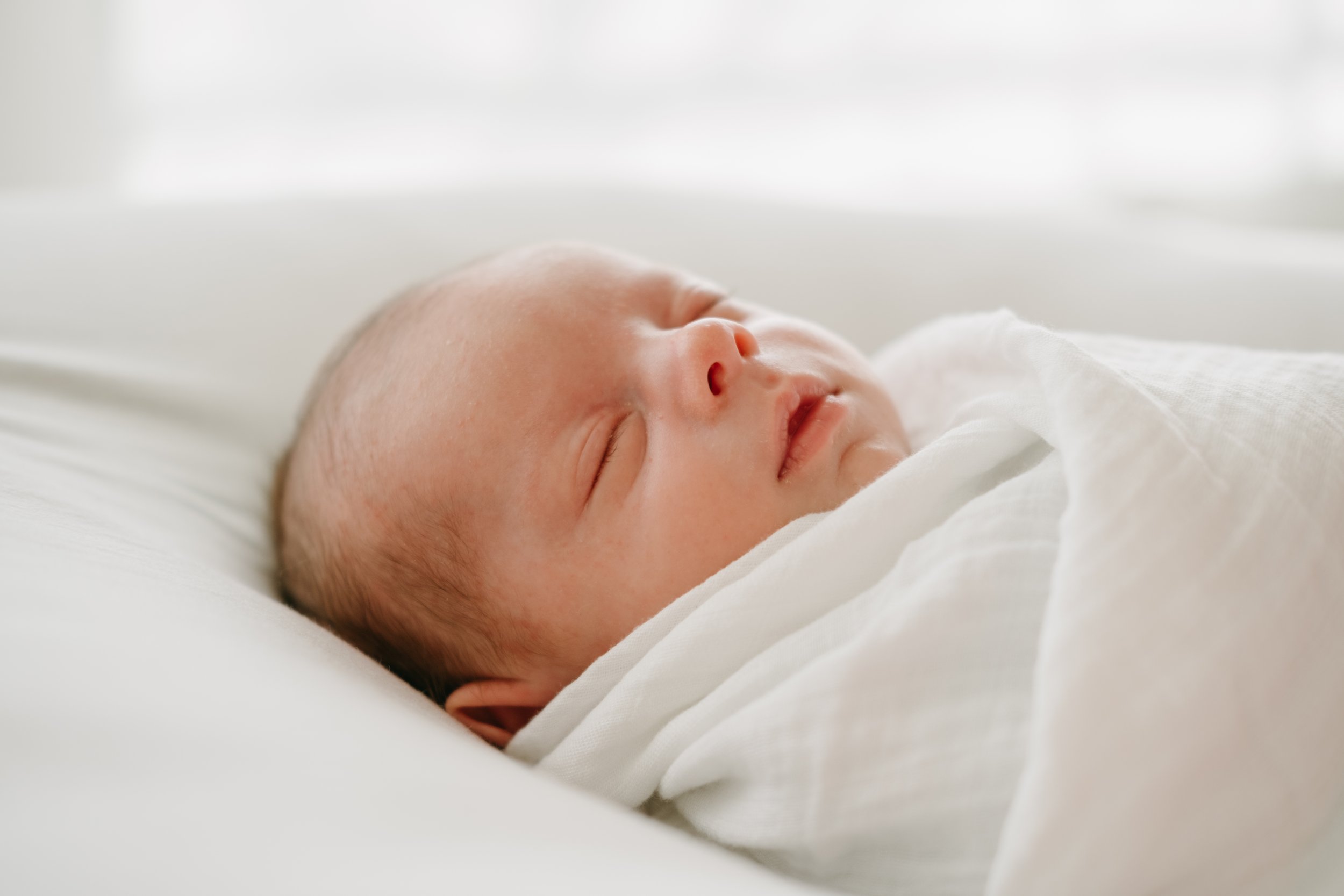 Close-up of a sleeping newborn baby wrapped in white fabric, resting on a soft white surface.
