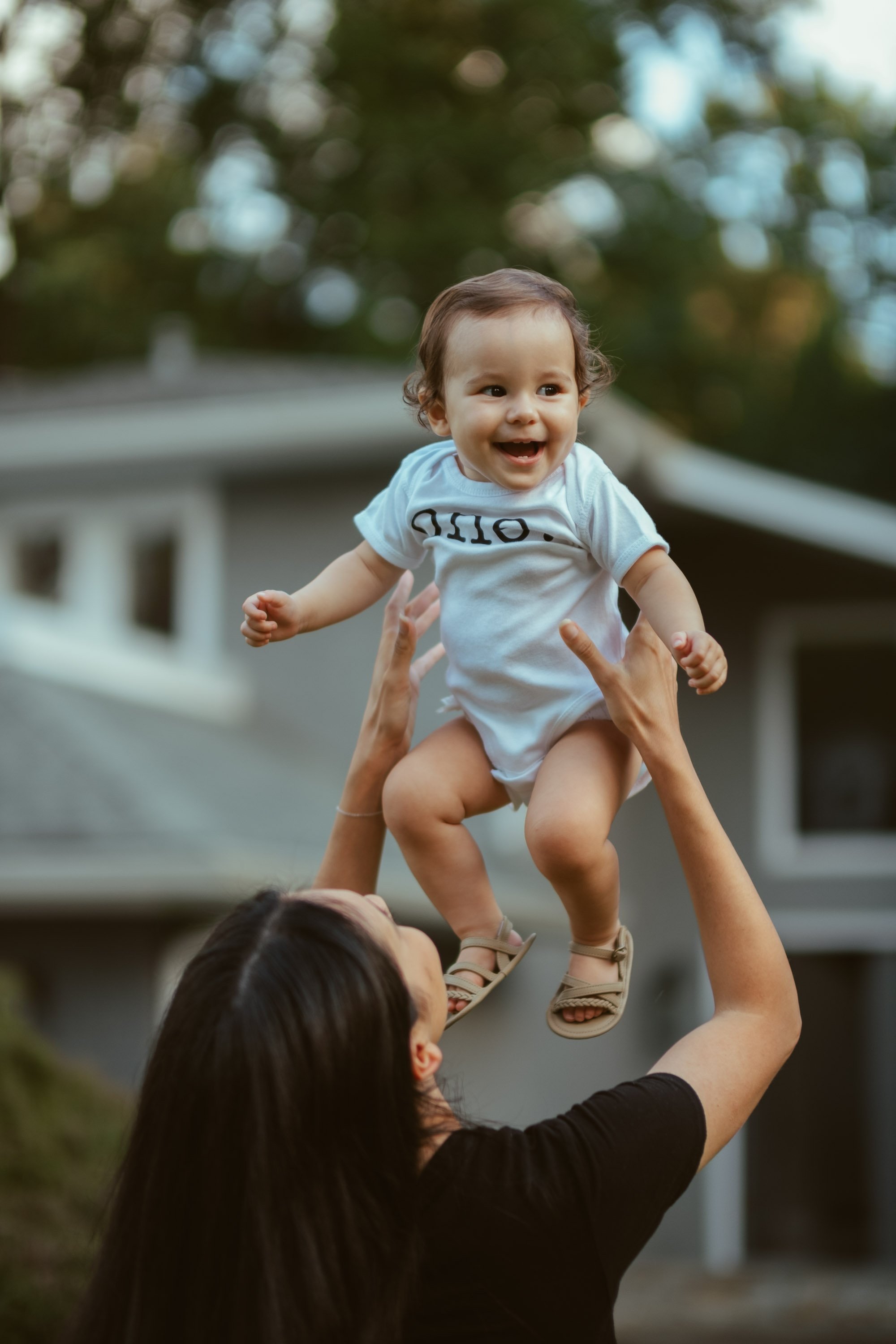 A smiling young child being lifted into the air by an adult outside in front of a house with trees in the background.