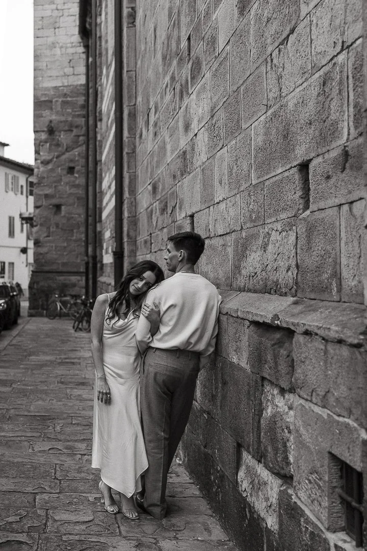 A couple in vintage attire leaning against a stone wall on a cobblestone street. The woman is in a satin dress and the man in a shirt and trousers. Black and white photograph.