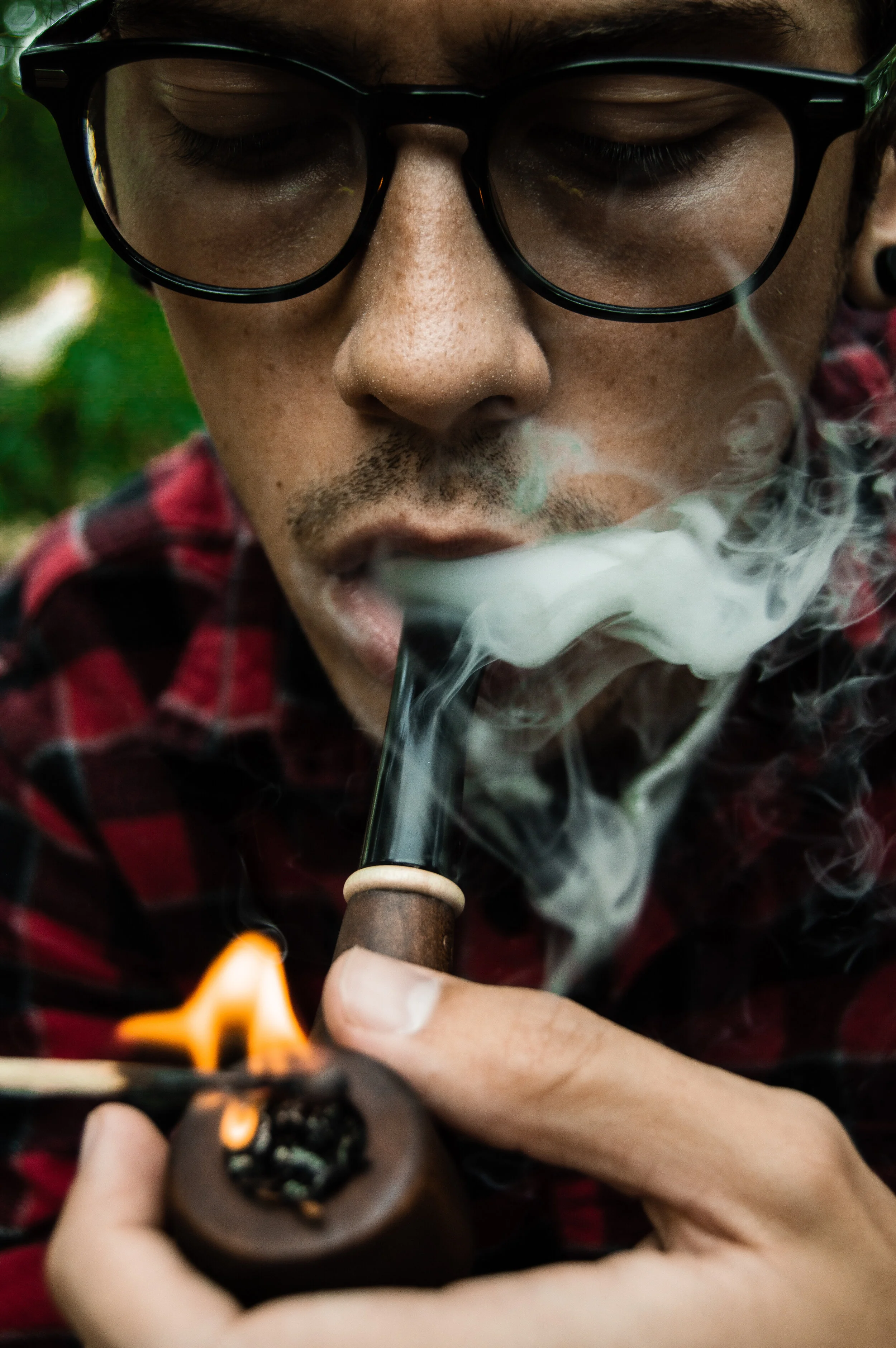 Close-up of a man with glasses smoking a pipe outdoors, with smoke swirling around his face.