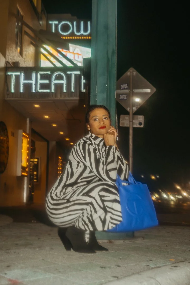 A woman with dark hair and red lipstick squats on the sidewalk near a building with a neon sign that reads 'TOW THEATRE'. She wears a zebra print coat, black boots, and holds a large blue handbag. It's nighttime with city lights in the background.