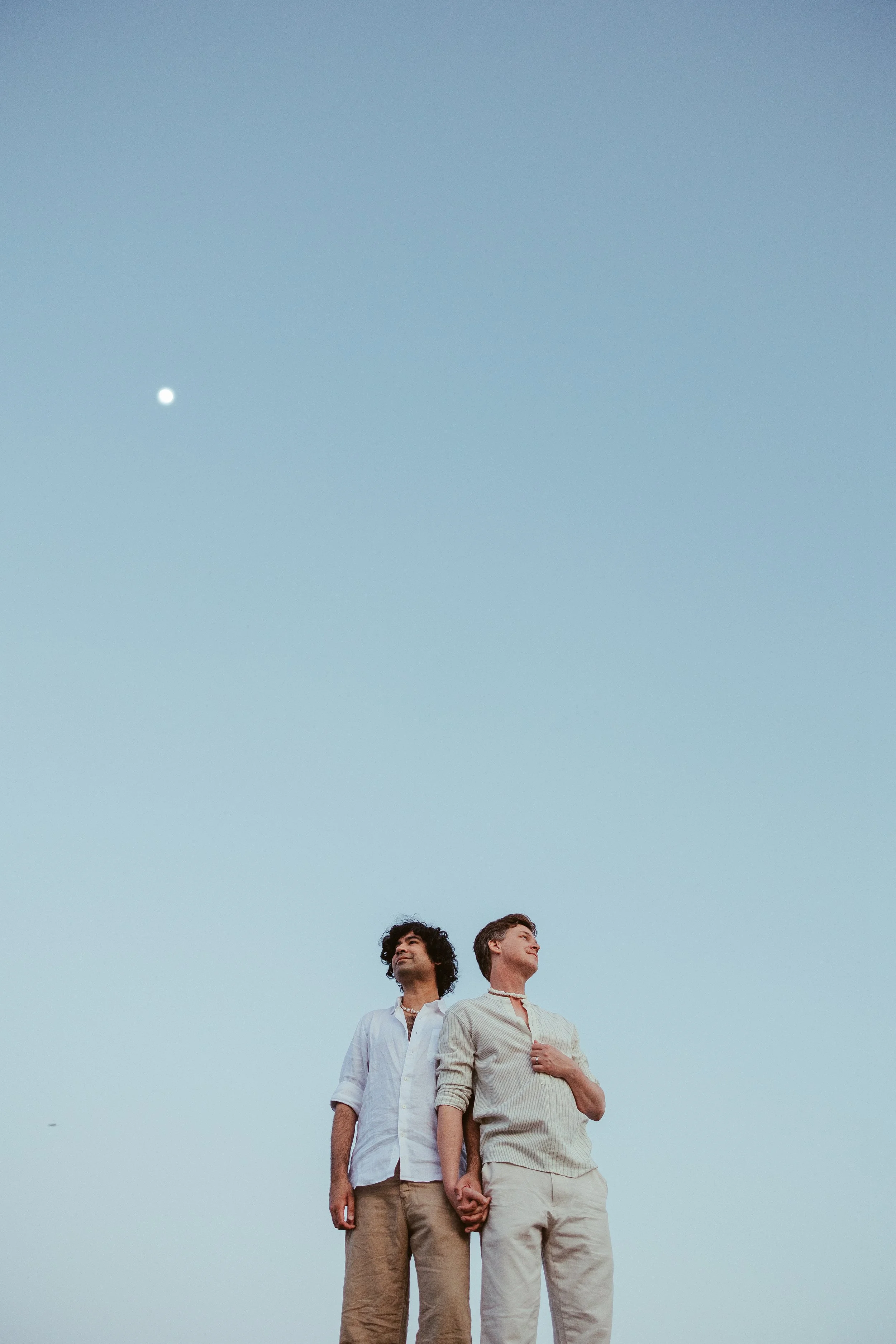 Two men holding hands and standing outdoors under a clear sky with the moon visible.