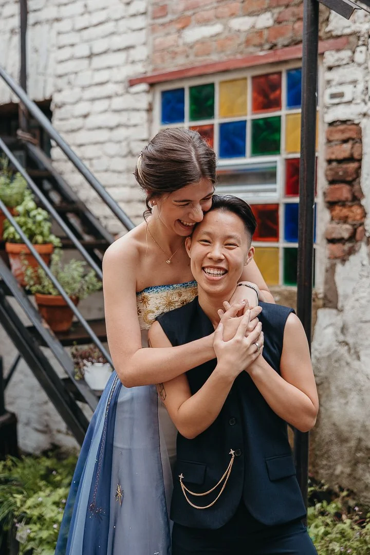 Two women smiling and hugging each other outdoors near a brick wall with a colorful stained glass window in the background. One woman is dressed in a strapless dress, and the other in a sleeveless blazer.