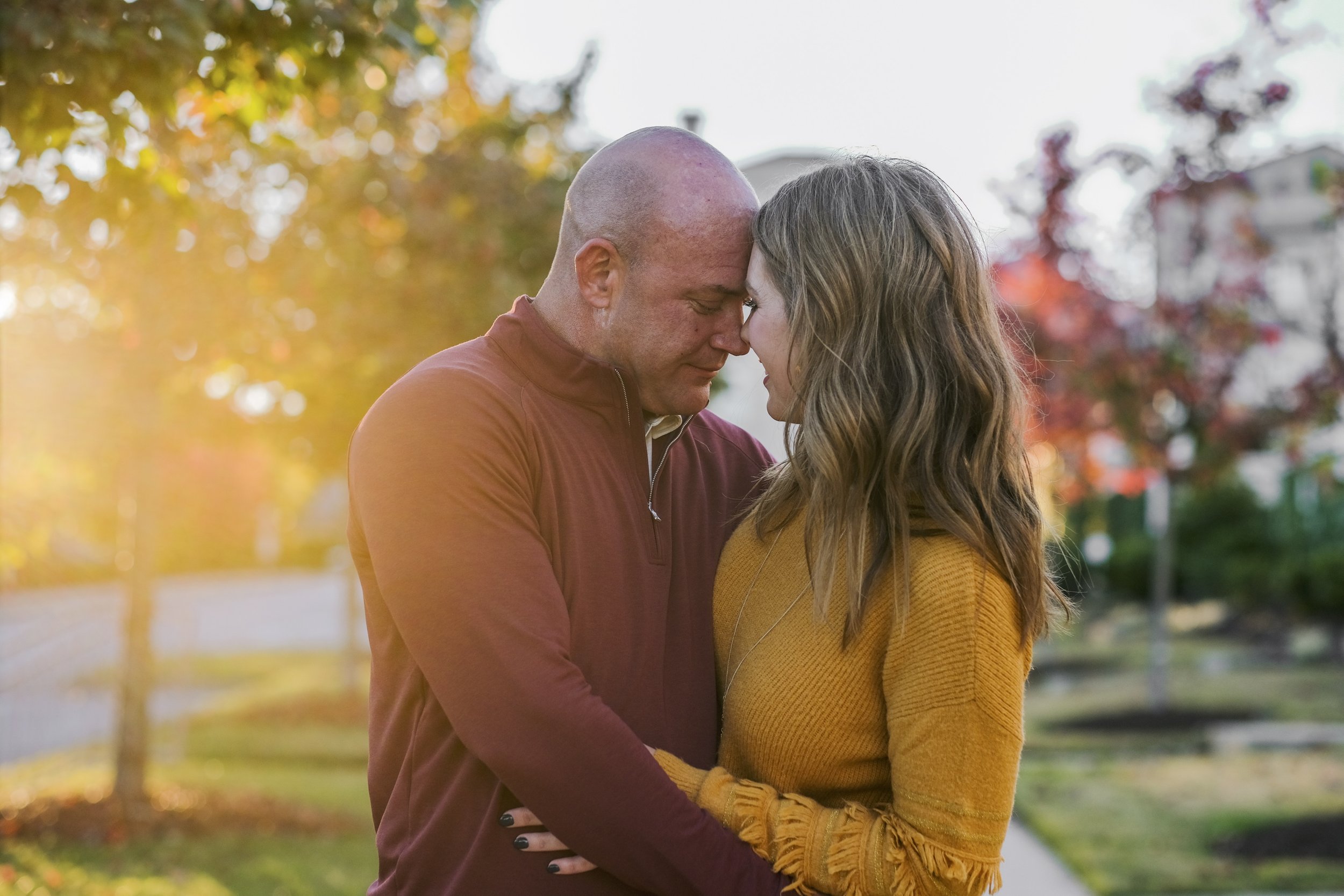A couple standing close together outdoors during fall, with their foreheads touching and eyes closed, in a tender moment.