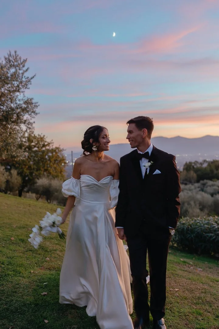 A bride and groom walking outdoors at sunset, holding hands, with the bride holding a bouquet of white flowers, in a scenic landscape with trees and distant mountains under a colorful sky with a visible moon.