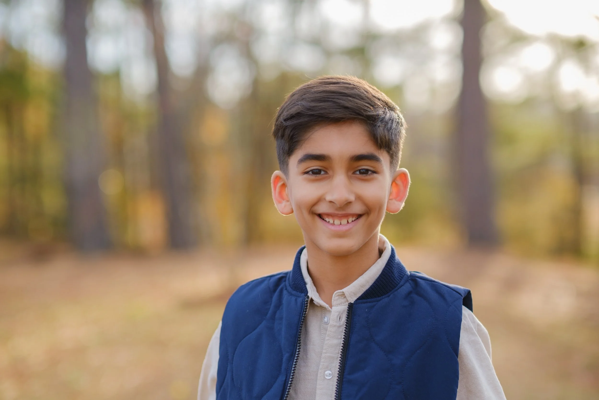 Smiling young boy outdoors in a forest with autumn leaves wearing a beige shirt and navy blue vest.
