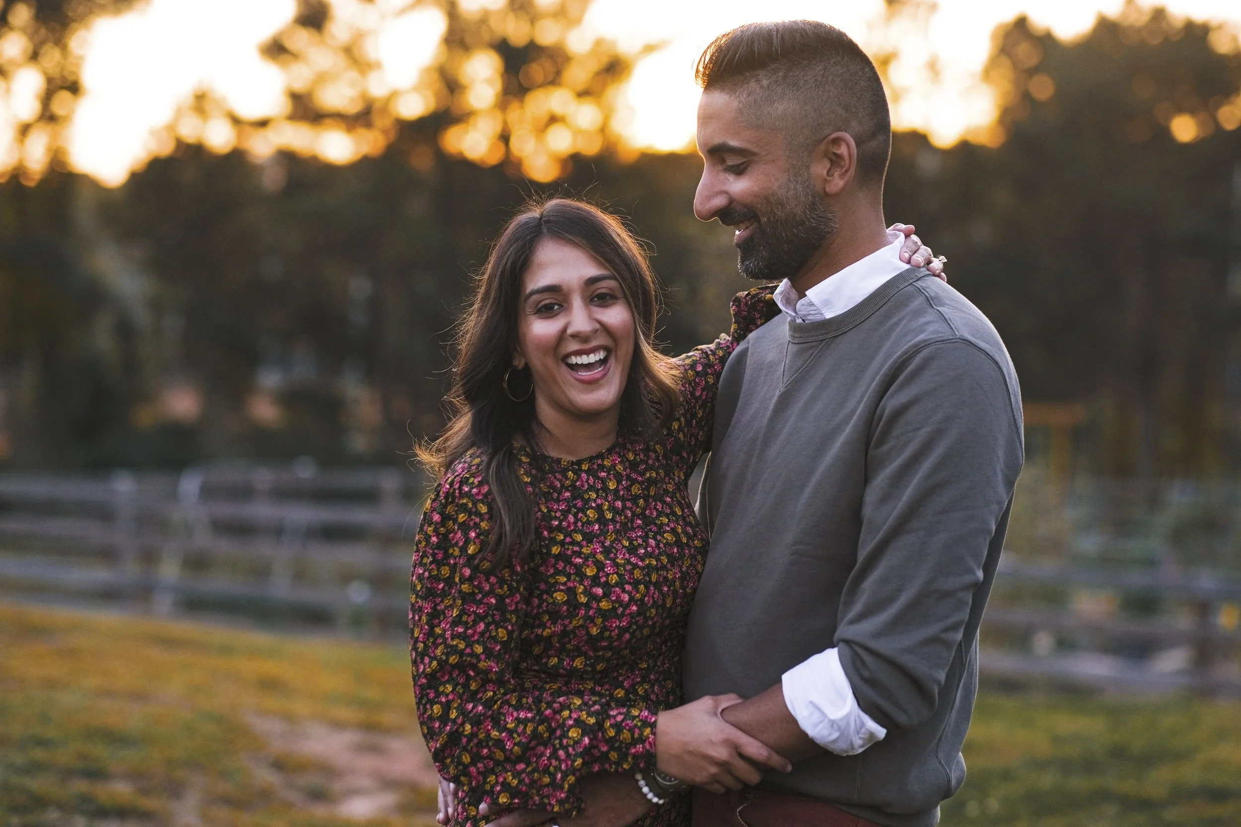 A happy couple outdoors at sunset, smiling and embracing each other, with a wooden fence and trees in the background.