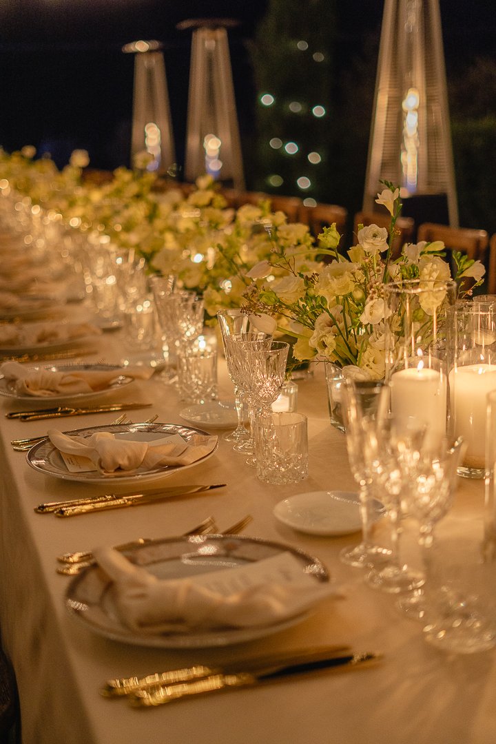 Elegant dinner table setup with white floral centerpiece, candles, and glassware, outdoors at night.