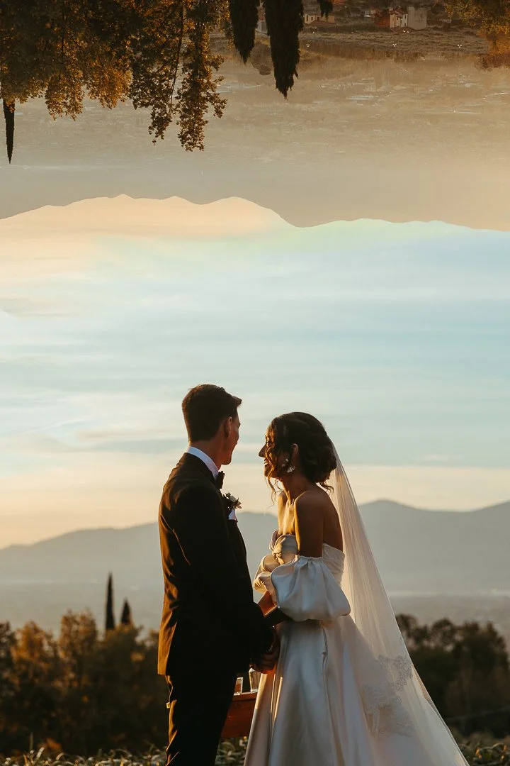 A bride and groom holding hands and gazing at each other during their wedding ceremony outdoors at sunset, with mountains in the background.