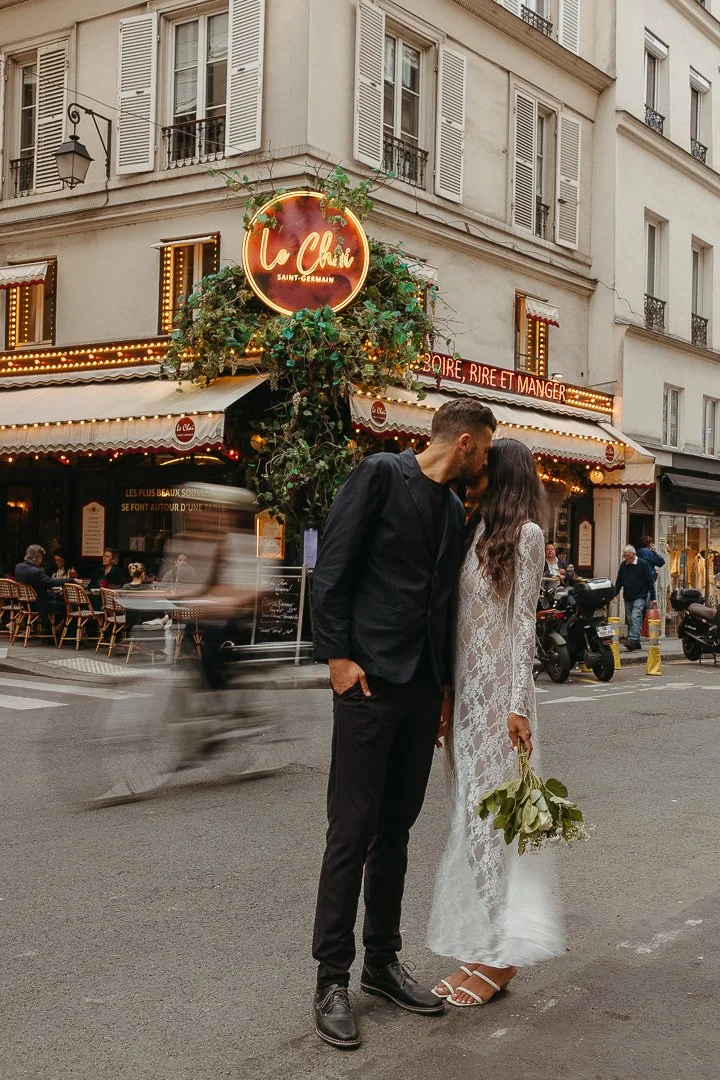 A couple dressed in wedding attire sharing a kiss in the street with a restaurant called La Chia in the background, decorated with lights and greenery, in a Parisian city setting.