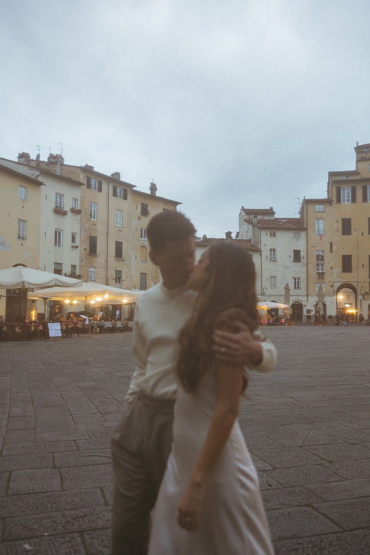 A couple kissing in a European town square during evening, surrounded by old buildings and outdoor restaurant seating with warm lights.