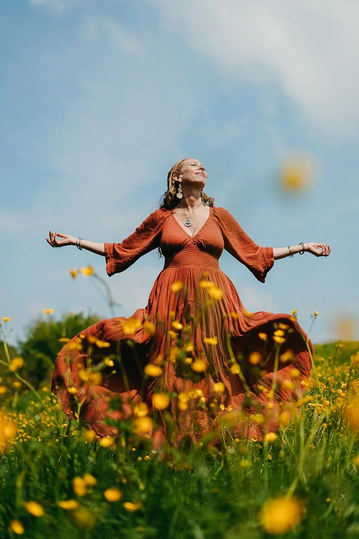 Woman in a long, rust-colored dress standing in a field of yellow flowers with her arms outstretched, looking upward with a joyful expression under a blue sky.