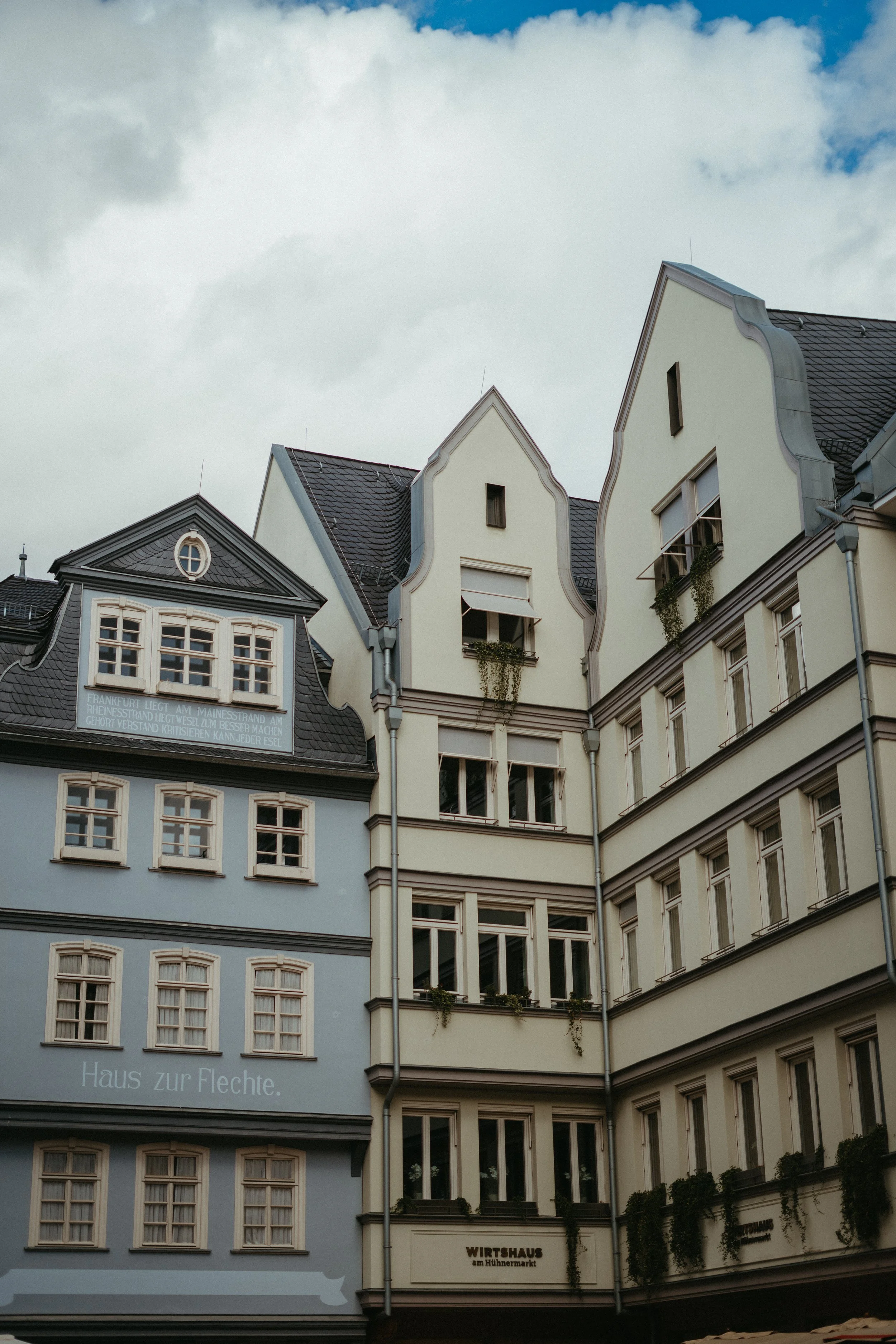 Multistory European-style building with decorative gable roofs and multiple windows. Some windows have plants, and there are signs in German, including "Haus zur Flechte" and "Wirtshaus am Hufenmarkt."