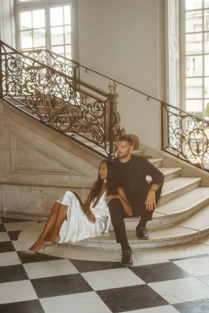 A man and woman sitting on marble stairs inside a grand building with large windows and ornate iron railing, the woman in a white dress and high heels, the man in black clothing and boots.