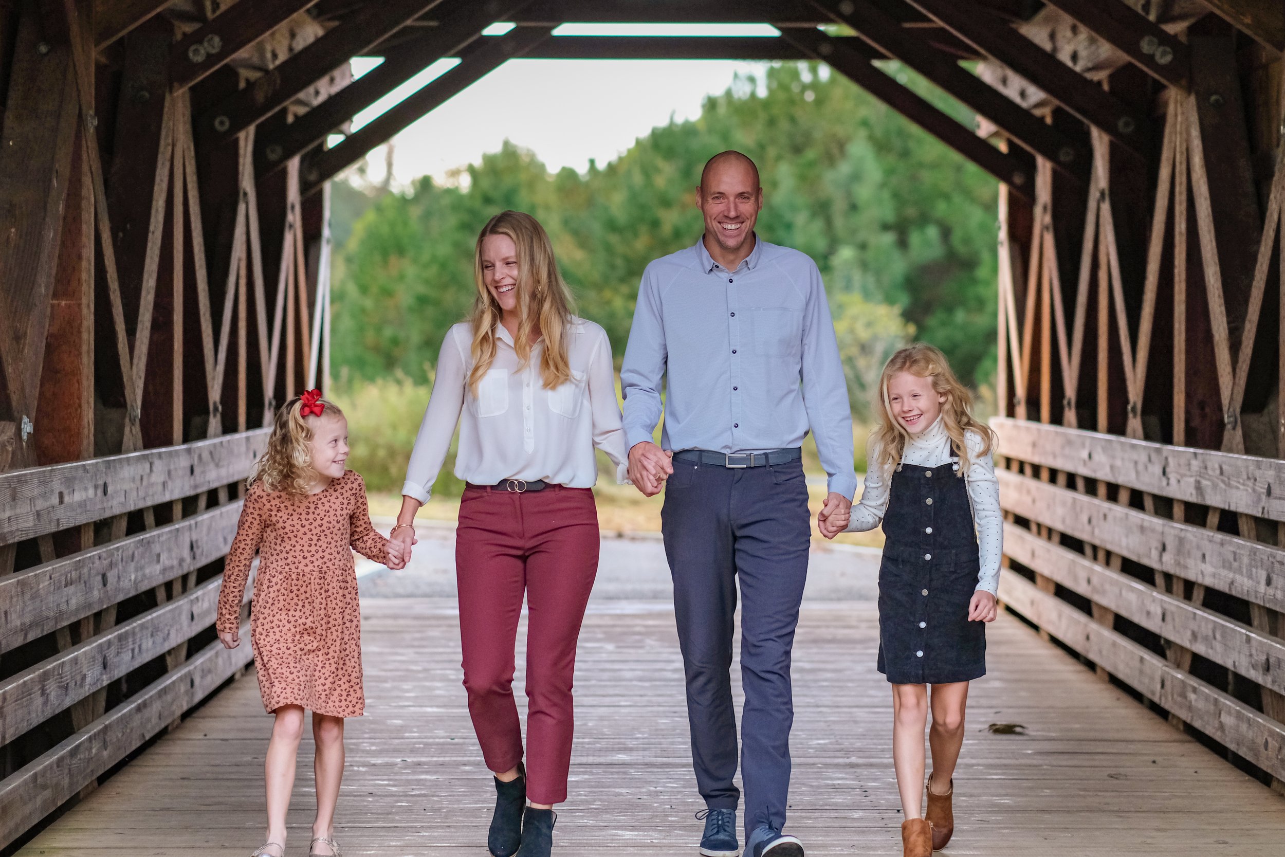 A family of four walking hand in hand on a wooden bridge surrounded by greenery.