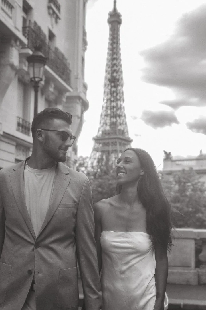 A man and woman walk hand in hand in front of the Eiffel Tower in Paris, France, smiling at each other. The photo is in black and white.