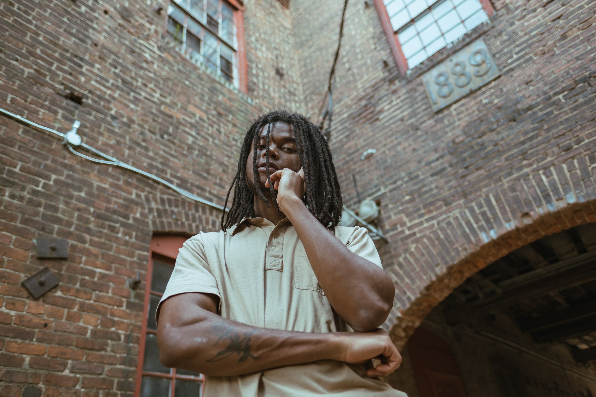 Young man with dreadlocks and tattoos leaning against brick wall in an alleyway, looking contemplative.