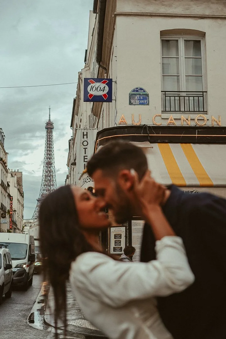 A romantic couple kissing on a street in Paris, with the Eiffel Tower visible in the background. The buildings have signs, including one that reads 'AU CANON' and a street sign indicating Rue Sully. The scene appears to be during an overcast day.