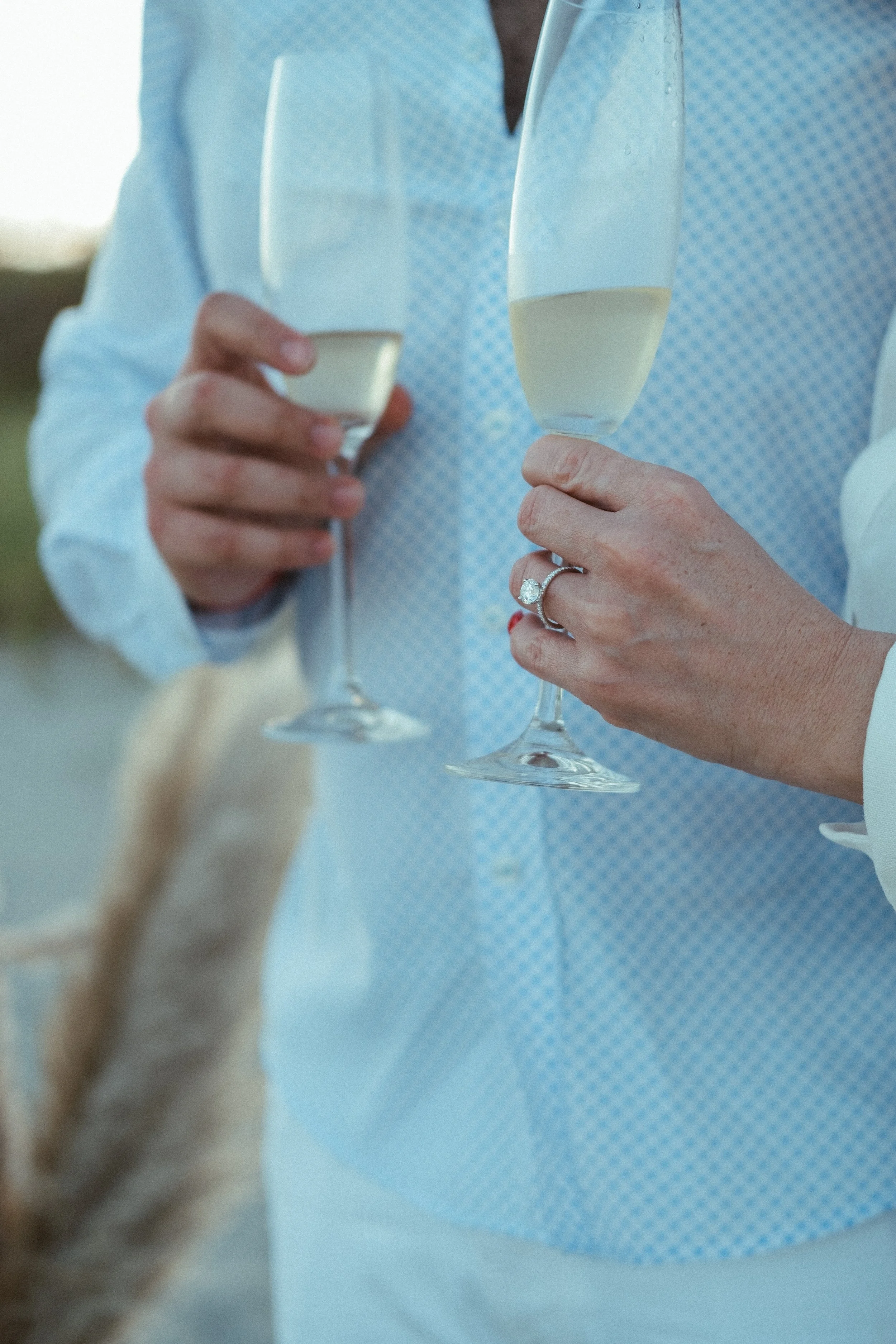 Two people toasting with glasses of champagne, one wearing a ring, in an outdoor setting.