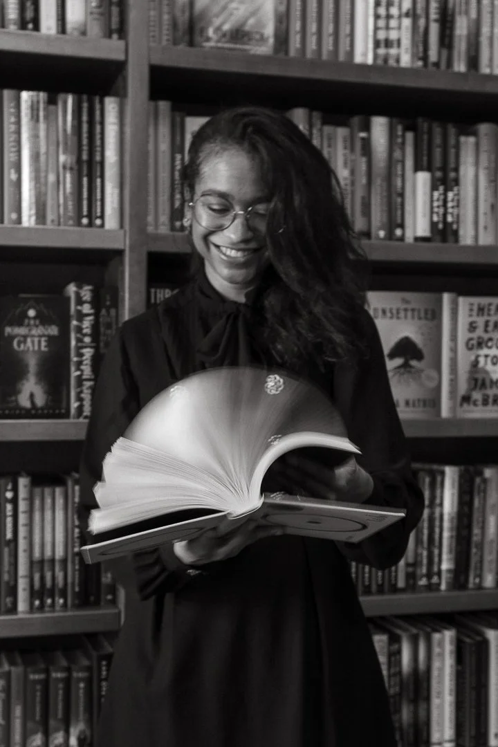 A woman with glasses and curly hair smiling while flipping through a book in a library with bookshelves in the background.