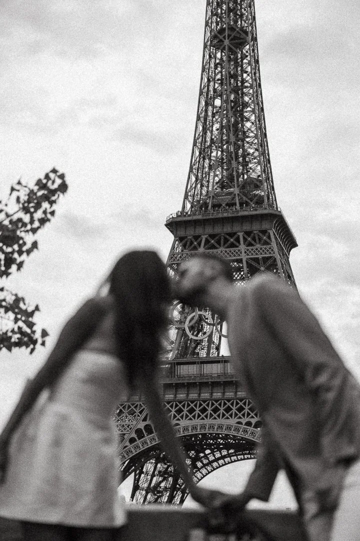 A couple kissing in front of the Eiffel Tower, with both people out of focus and the tower sharply focused in black and white.