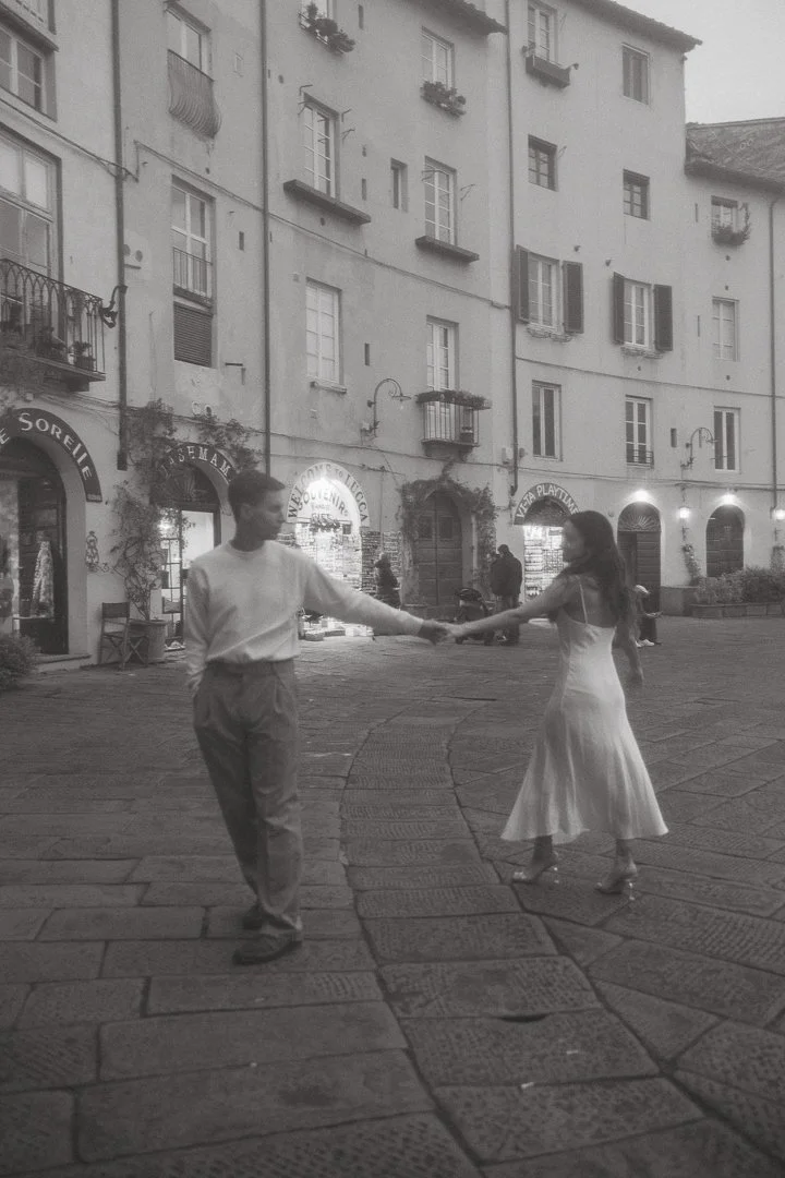 A young man and woman dancing and holding hands on a European city street at night, with historic buildings and shop signs in the background.