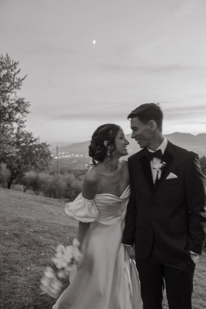 A black and white photo of a bride and groom smiling at each other outdoors during sunset, with mountains and trees in the background.
