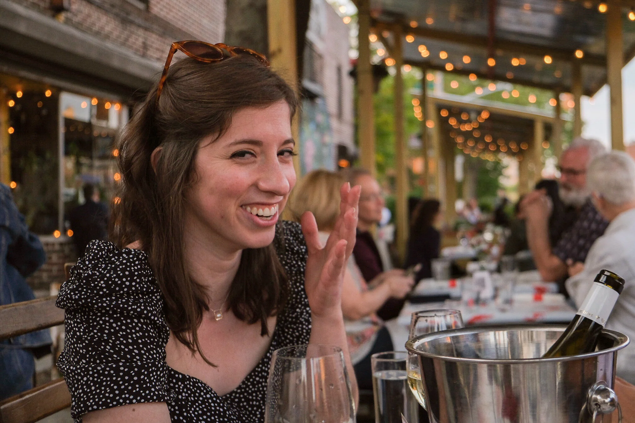 A smiling woman with brown hair, wearing sunglasses on her head and a black and white polka dot top, sitting at a table outdoors during evening, with a glass of white wine in front of her and an ice bucket with a bottle of wine. Other people are dini