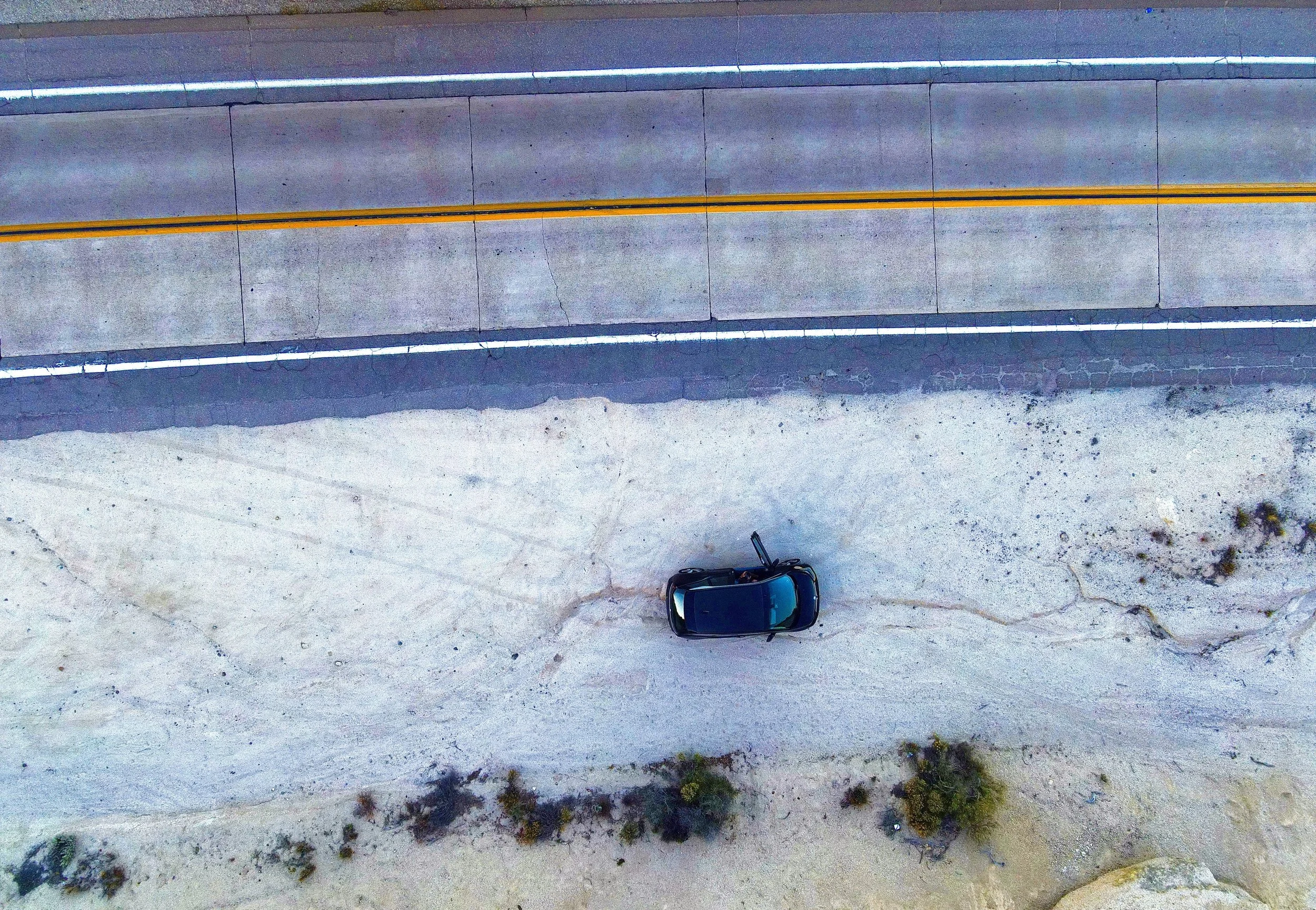 An aerial view of a sandy outdoor area with a single dark-colored car parked near a concrete curb or wall. Two parallel white lines are painted on the paved surface above the concrete, possibly indicating a boundary or lane markings.