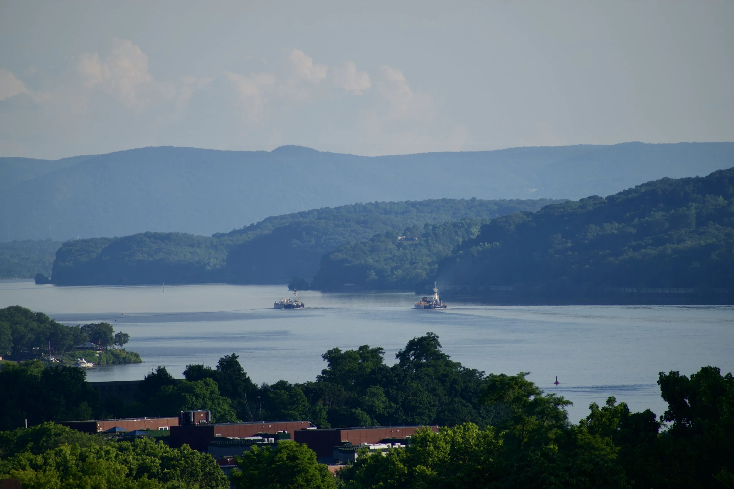 A scenic view of a large river with two boats sailing on it, surrounded by lush green trees and rolling hills in the background under a partly cloudy sky.