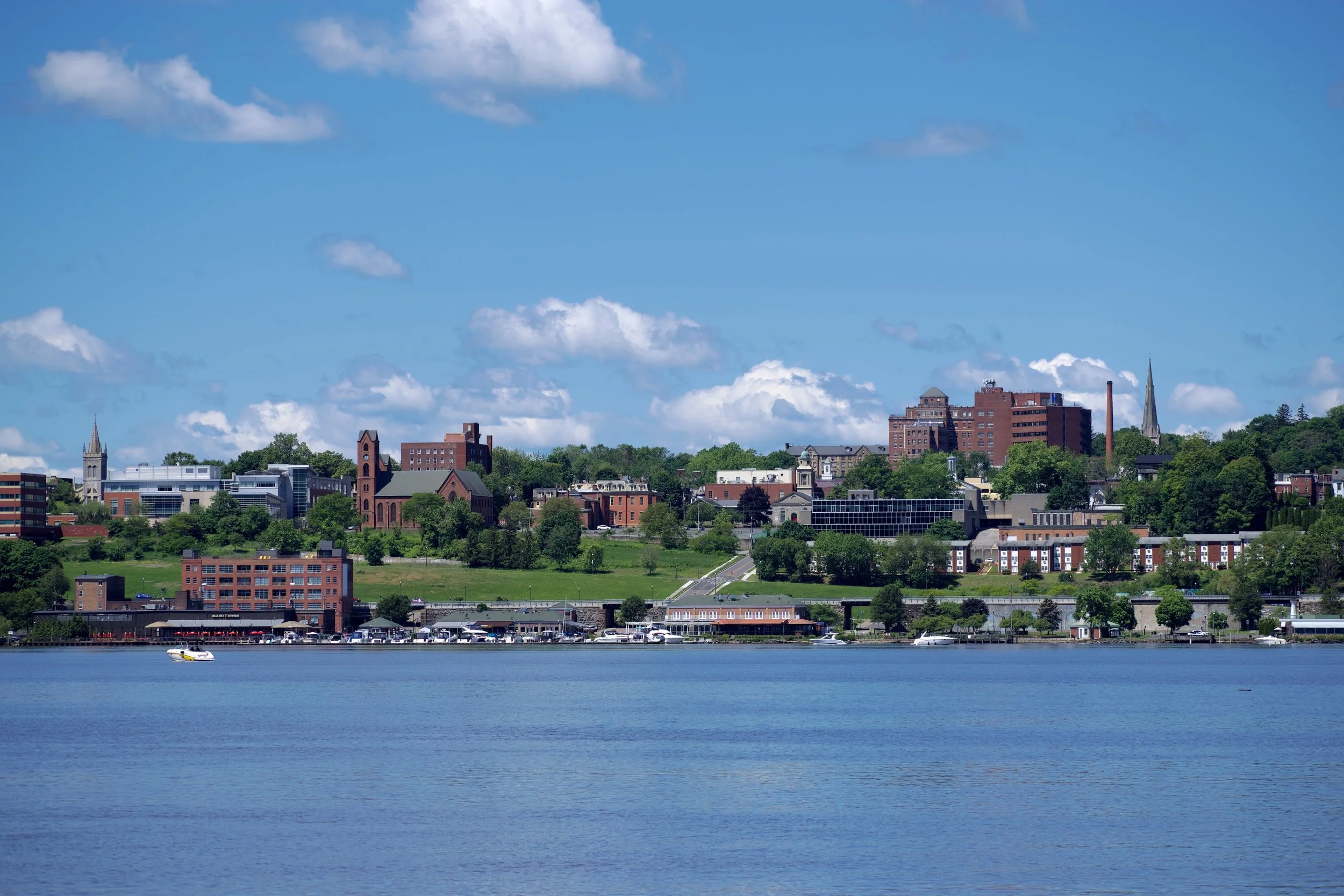 A city skyline along a river with several buildings, churches, and green trees on a clear day with blue sky and white clouds.