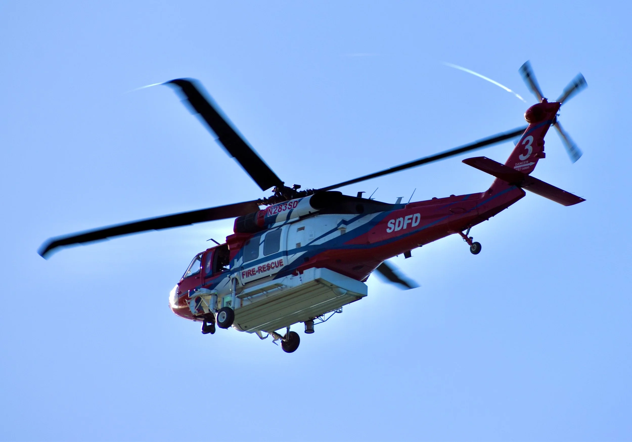 A red and white firefighting helicopter with a rescue platform beneath it, flying against a clear blue sky.