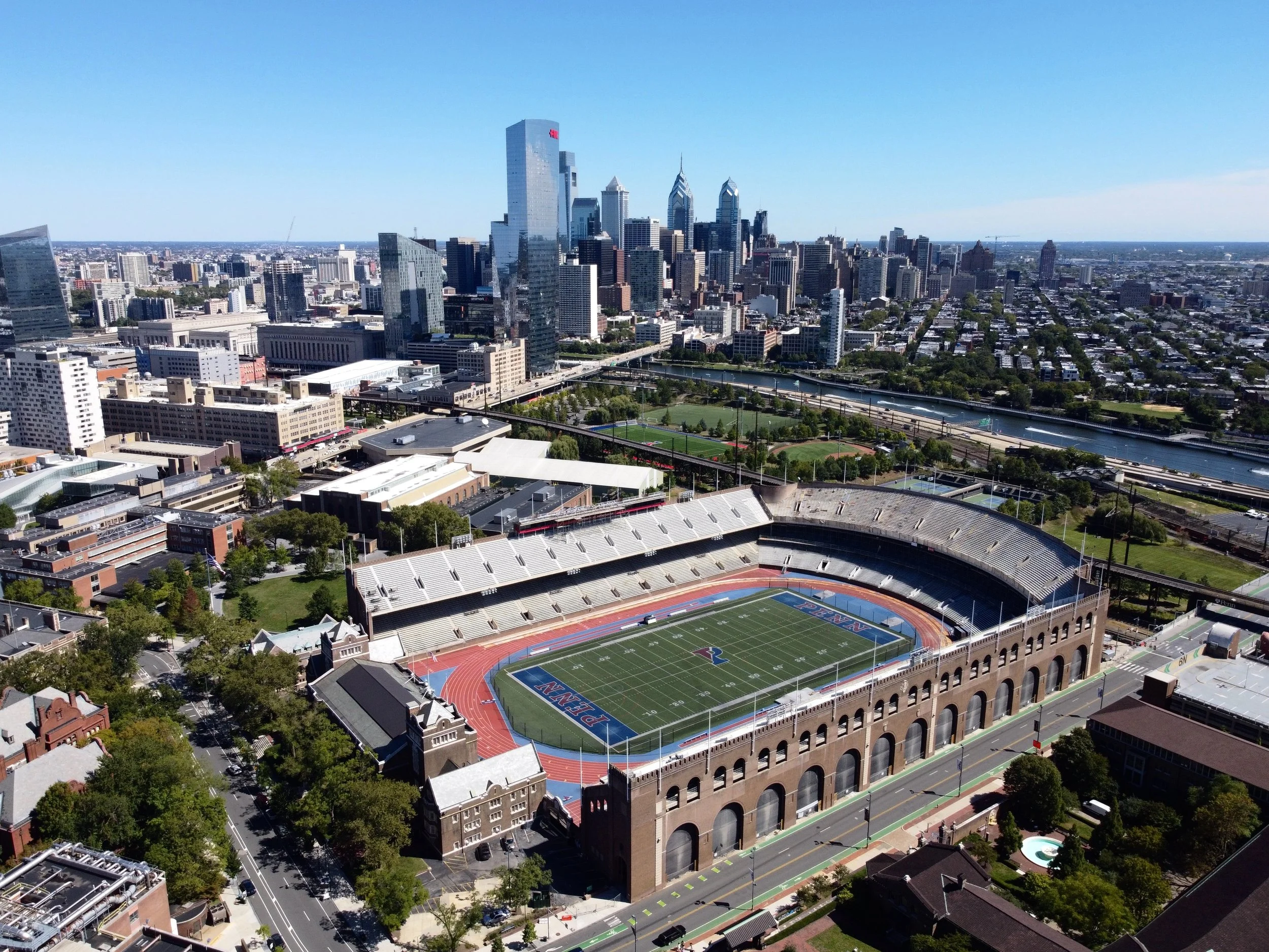 Aerial view of a city skyline with high-rise buildings, a football stadium with a track, and a river winding through the city under a clear blue sky.