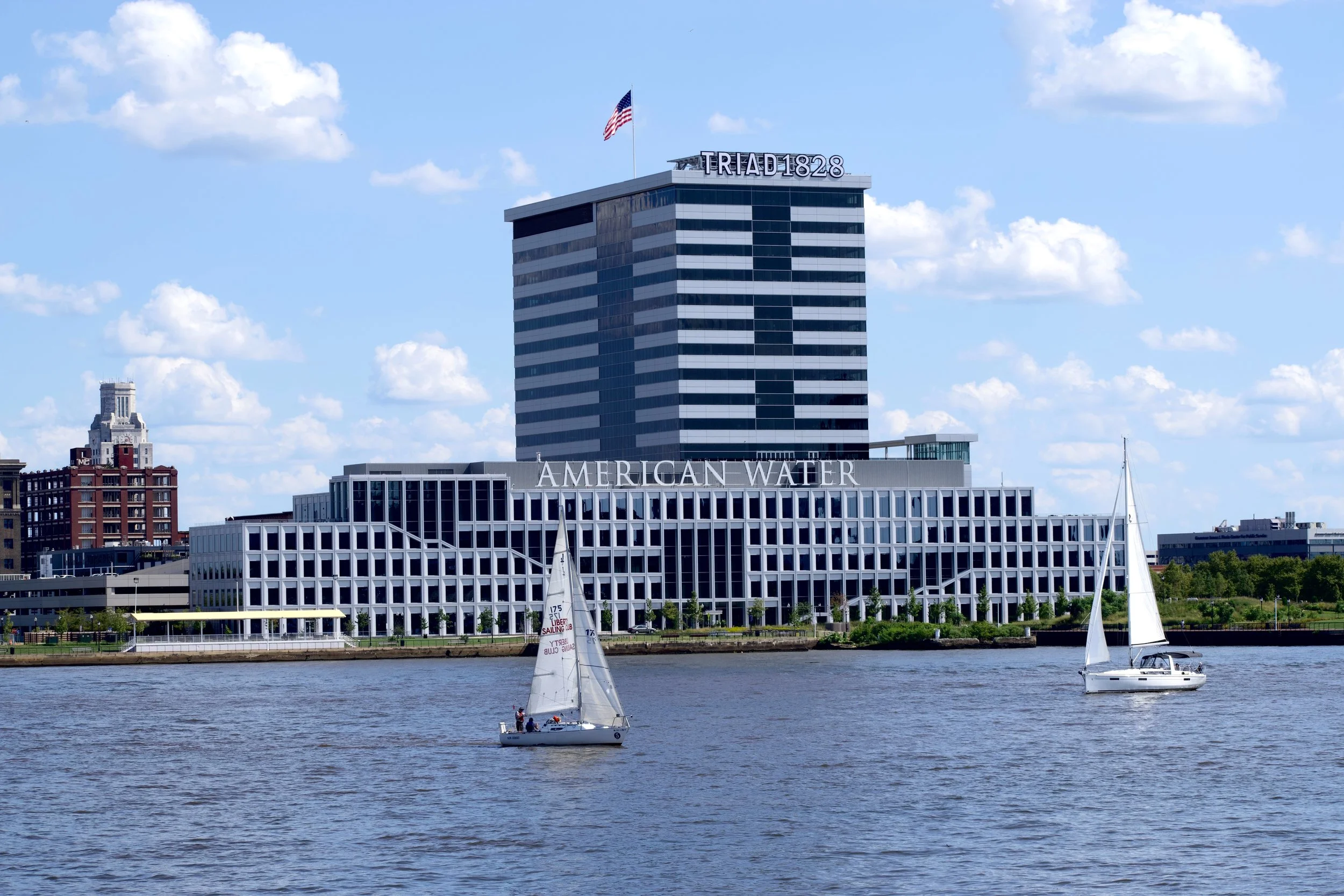 A cityscape view featuring a tall modern building with the word 'TRIAD 1828' on top, an American flag, a waterfront, and sailboats on the water with blue skies and scattered clouds in the background.