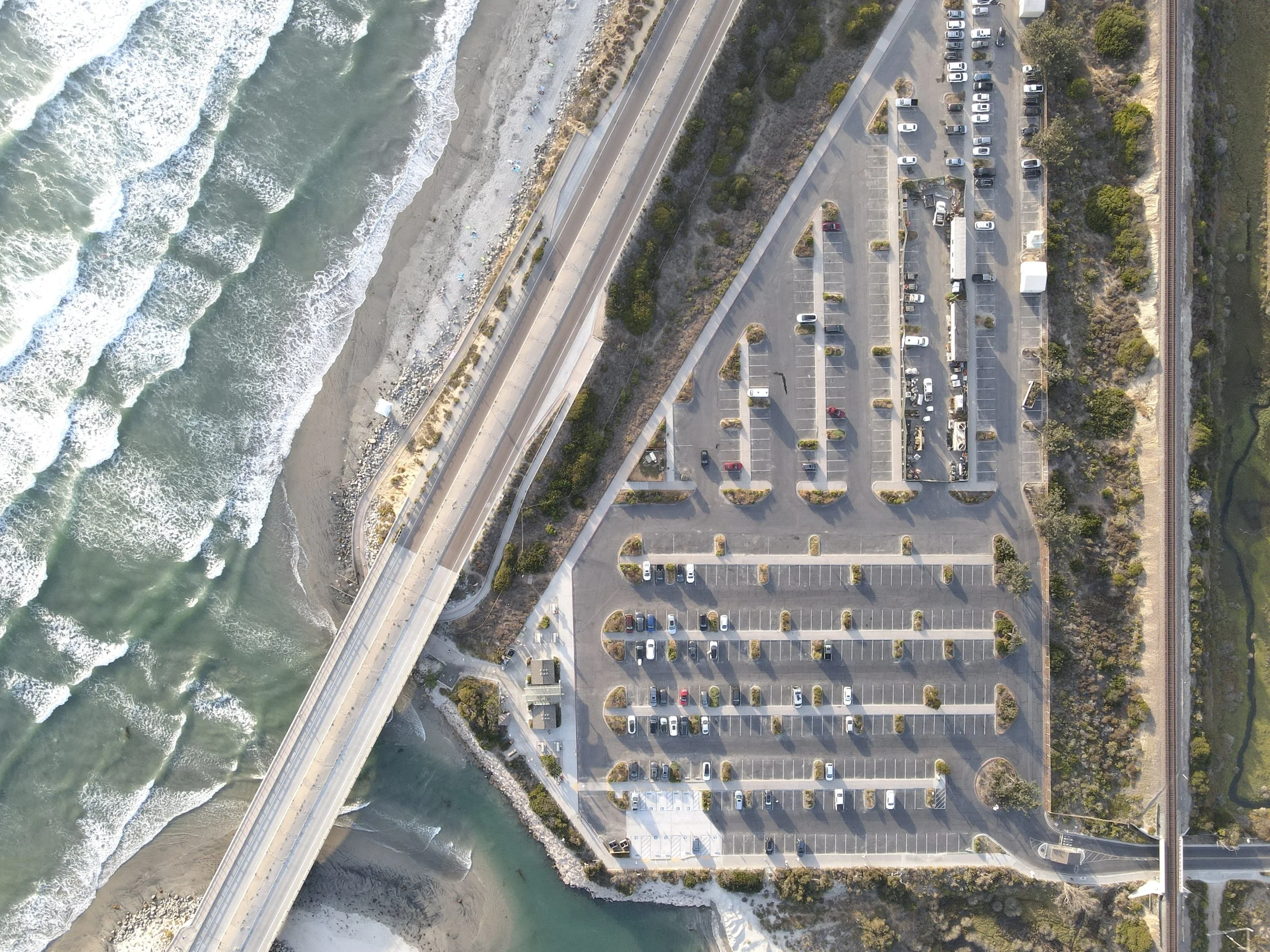 An aerial view of a parking lot adjacent to the ocean, with a bridge crossing over connecting the parking area to the beach and ocean waves.