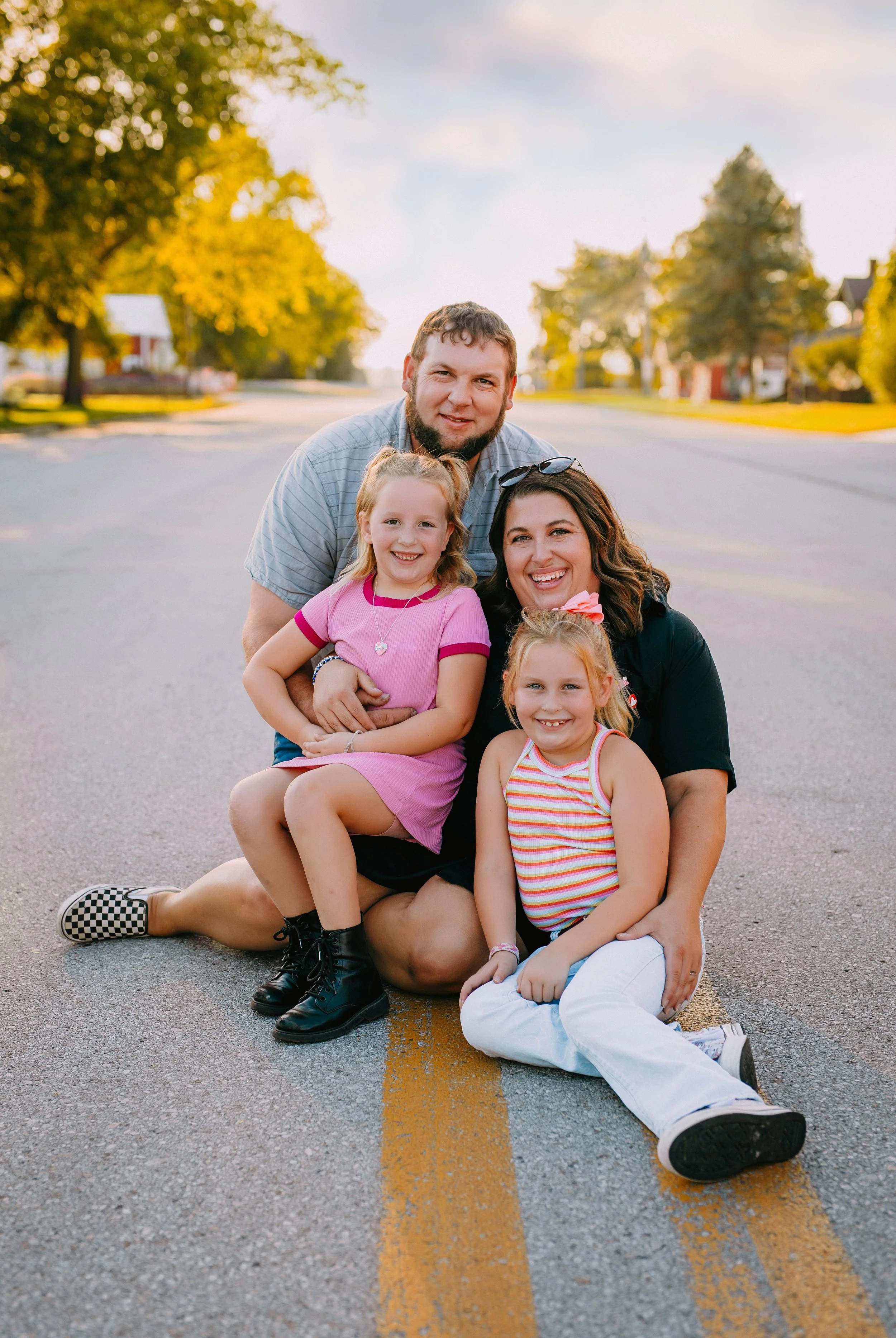 A family of five sitting and kneeling on a city street with trees and houses in the background during sunset, smiling at the camera.