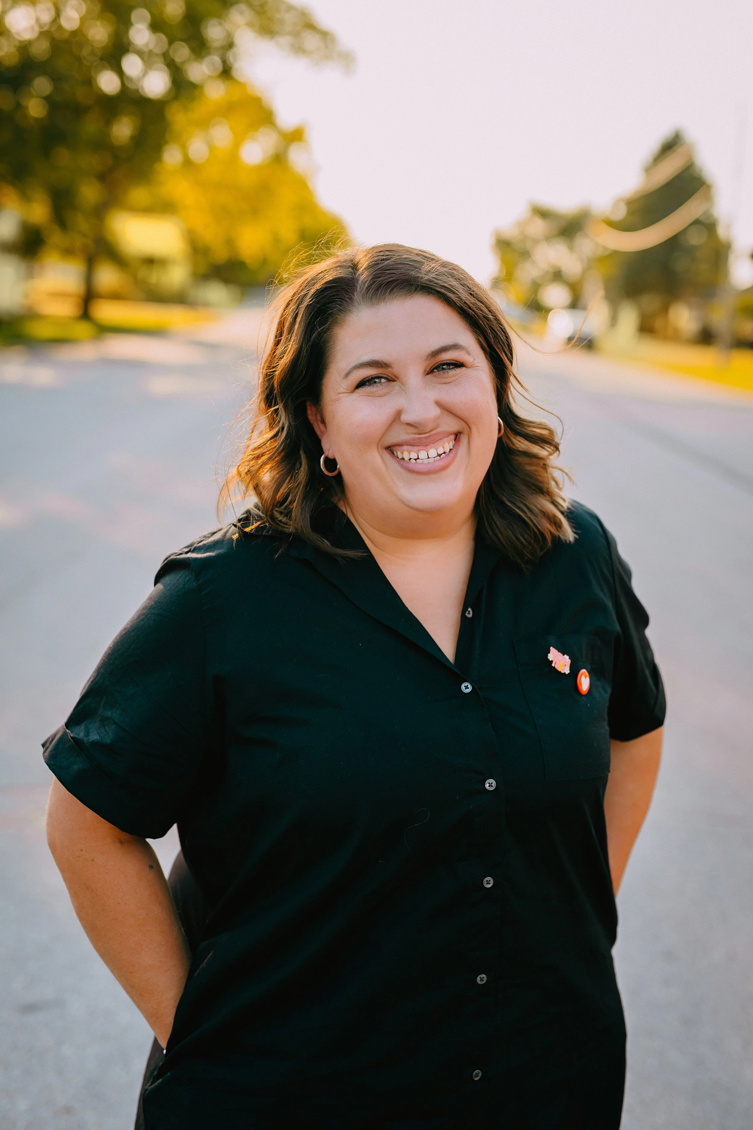 Maggie Allen smiling outdoors during sunset, wearing a black shirt with pins, standing on a street lined with trees and houses.
