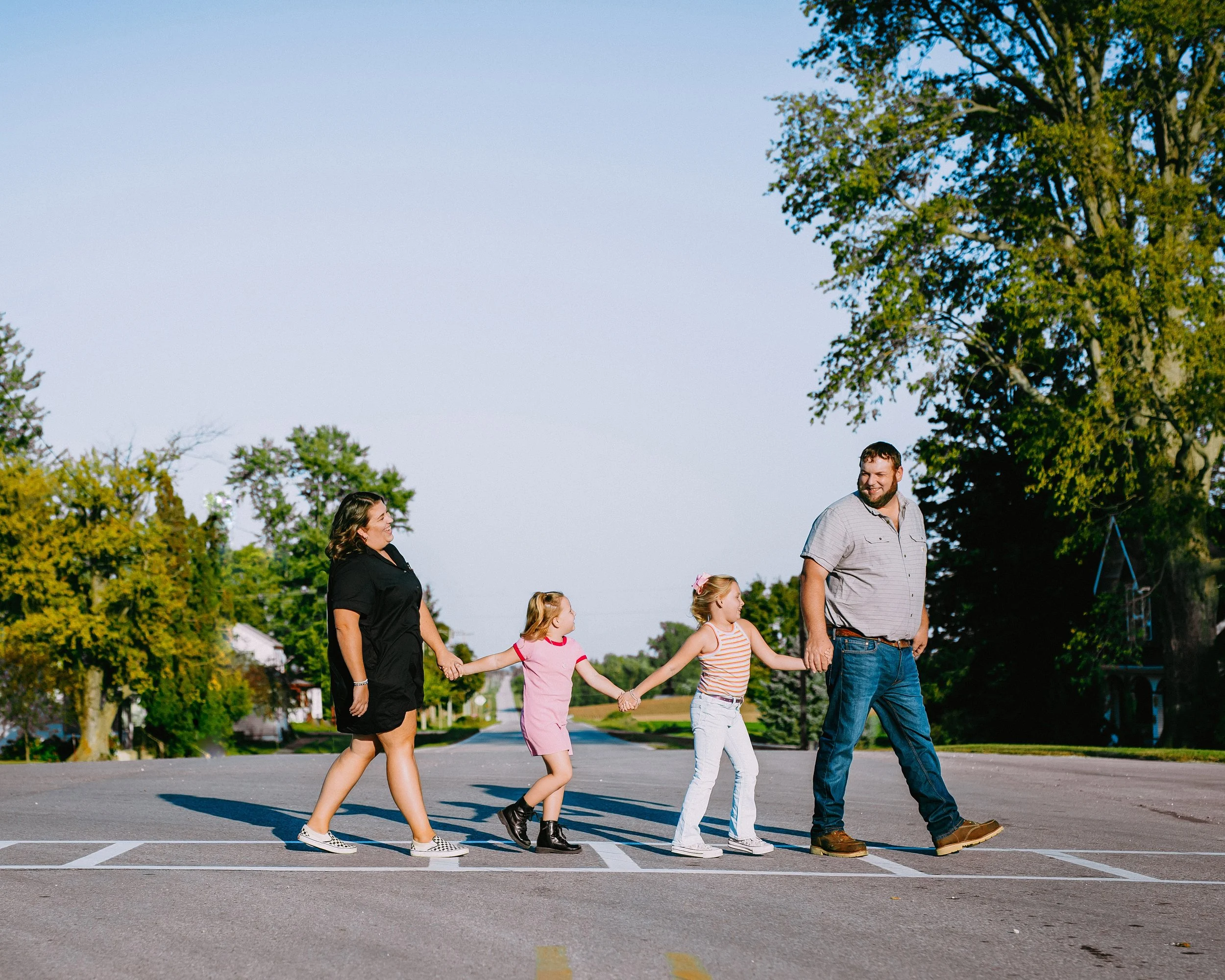 A family walking hand in hand across a crosswalk outdoors on a sunny day, with trees and a clear blue sky in the background.