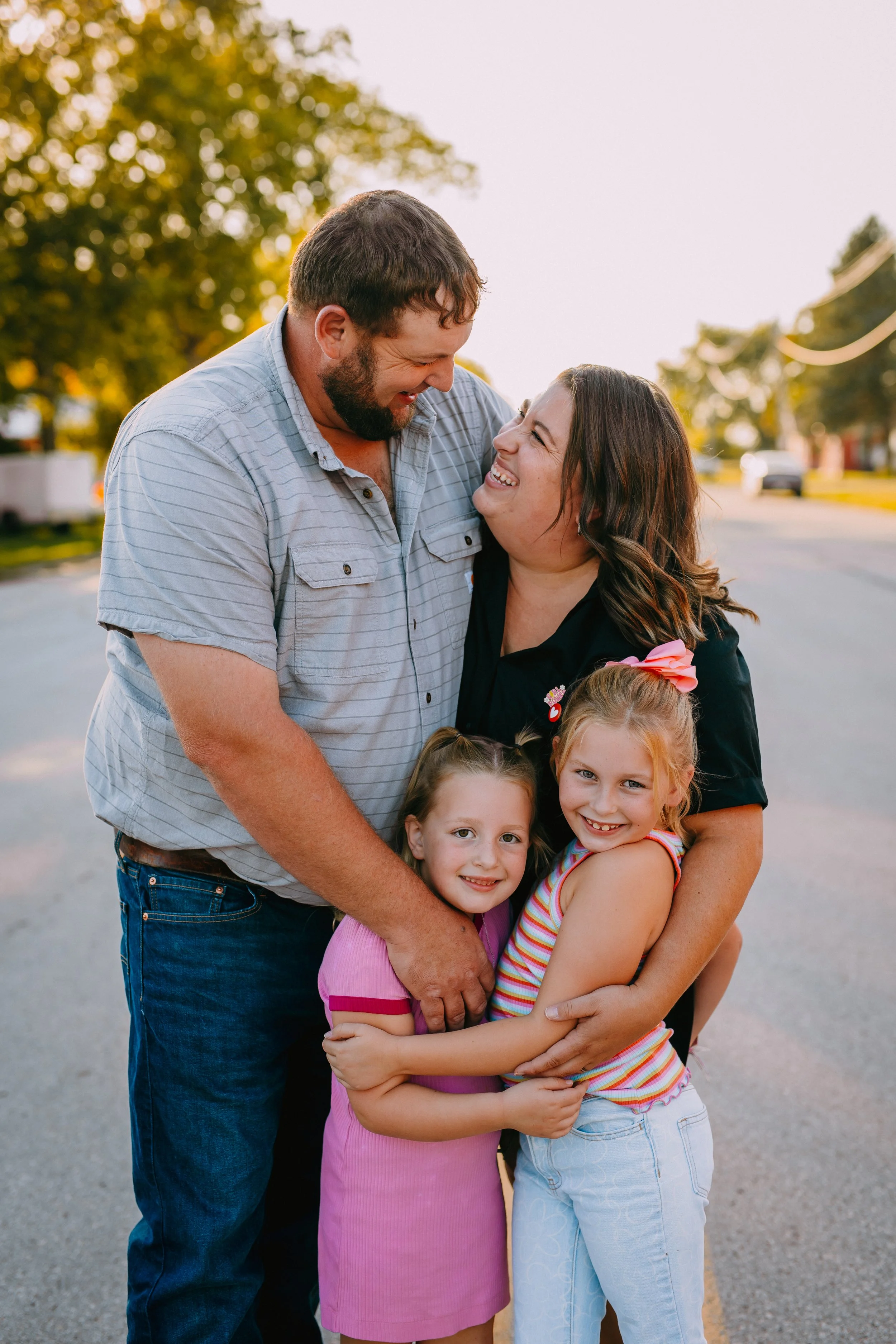 A family of four with a man, woman, and two young girls, sharing a joyful embrace outdoors during daylight.
