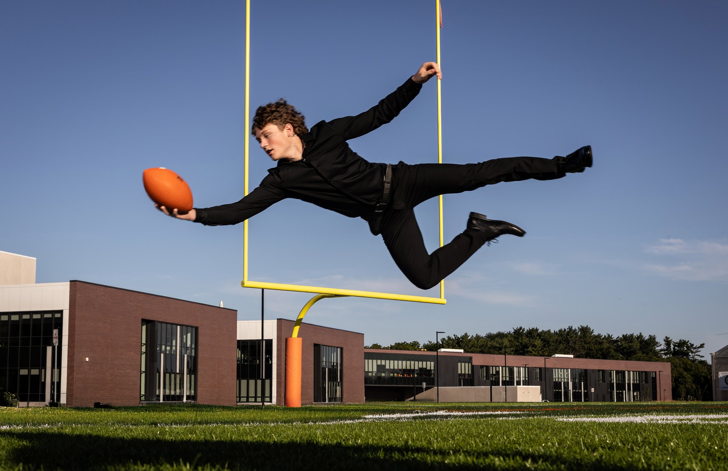 A man in black clothing is flying horizontally in the air above a football field, holding a football in his right hand, with a goalpost behind him and buildings in the background.