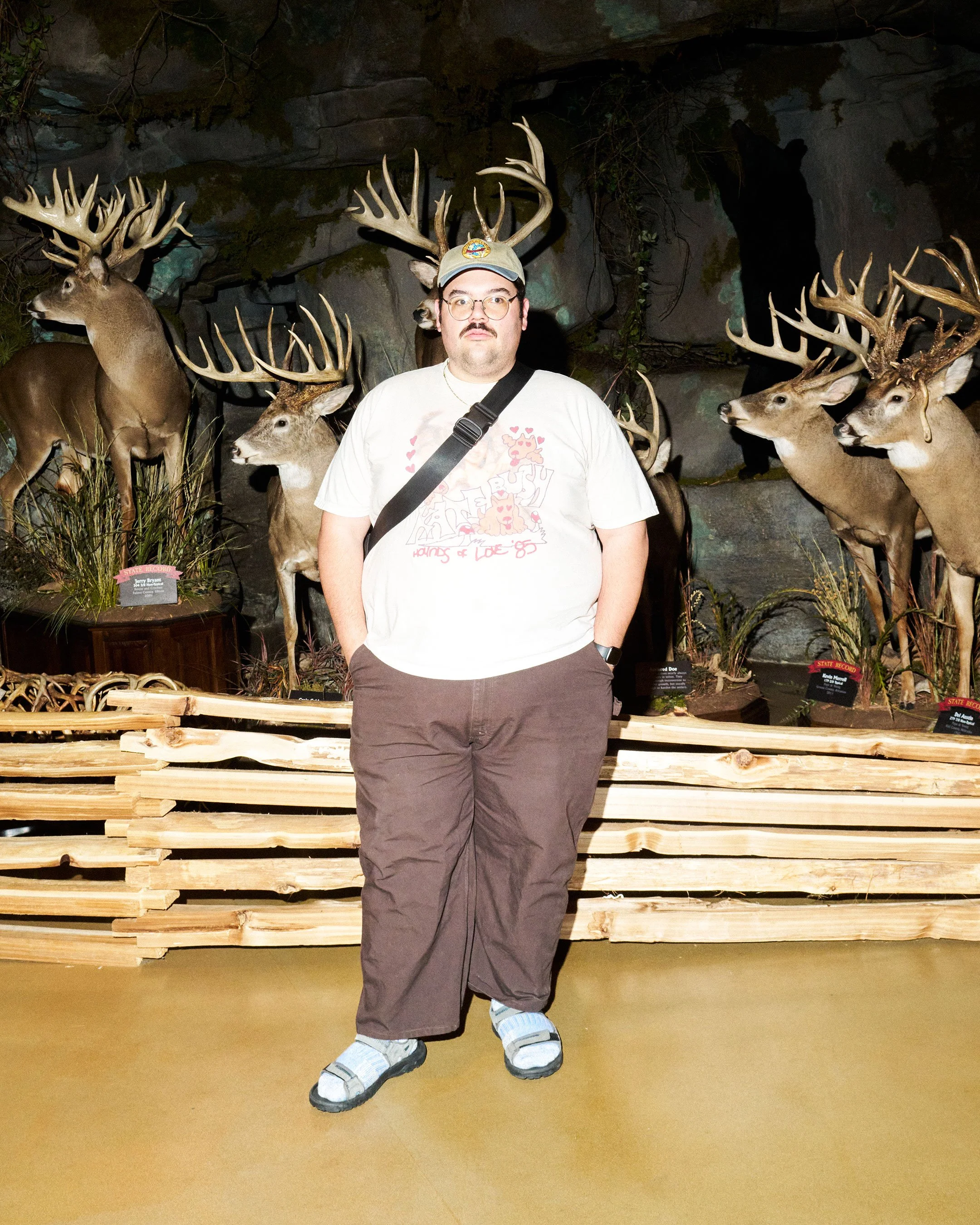 A man wearing glasses, a baseball cap, a white T-shirt with pink hearts, brown pants, and white sandals stands in front of a display of mounted deer heads with large antlers inside a museum or exhibit.